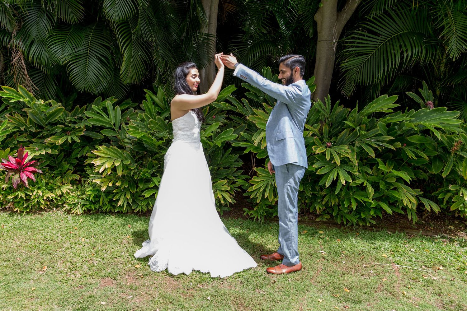 A bride and groom dance outdoors in a lush tropical garden, she in a white gown, he in a light blue suit.