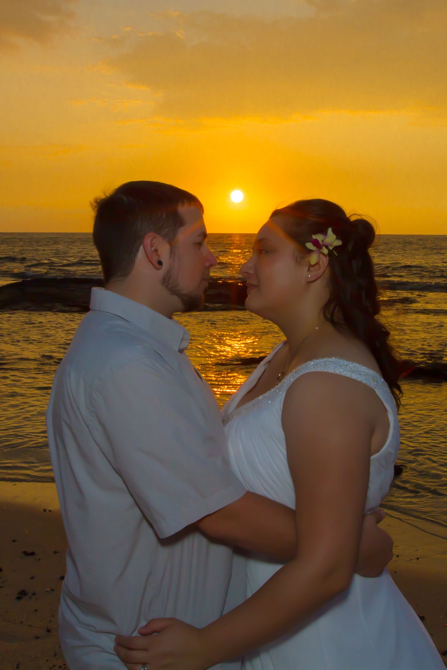 A couple embraces on a Kikaua beach, in Kona at sunset as golden sunlight reflects off the ocean waves behind them.