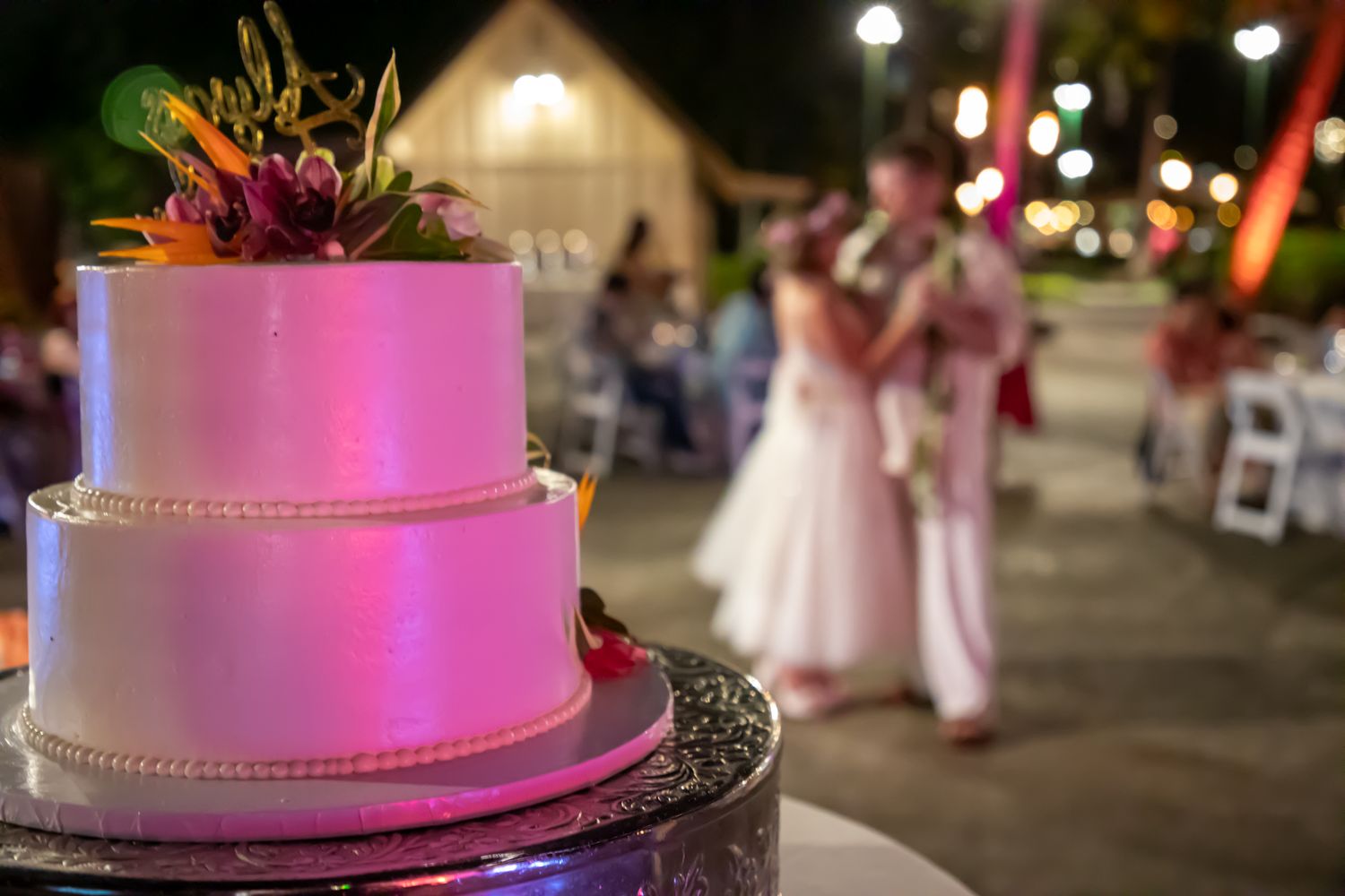 Two-tier white wedding cake with floral topper glowing pink, with couple dancing at outdoor evening reception in background.