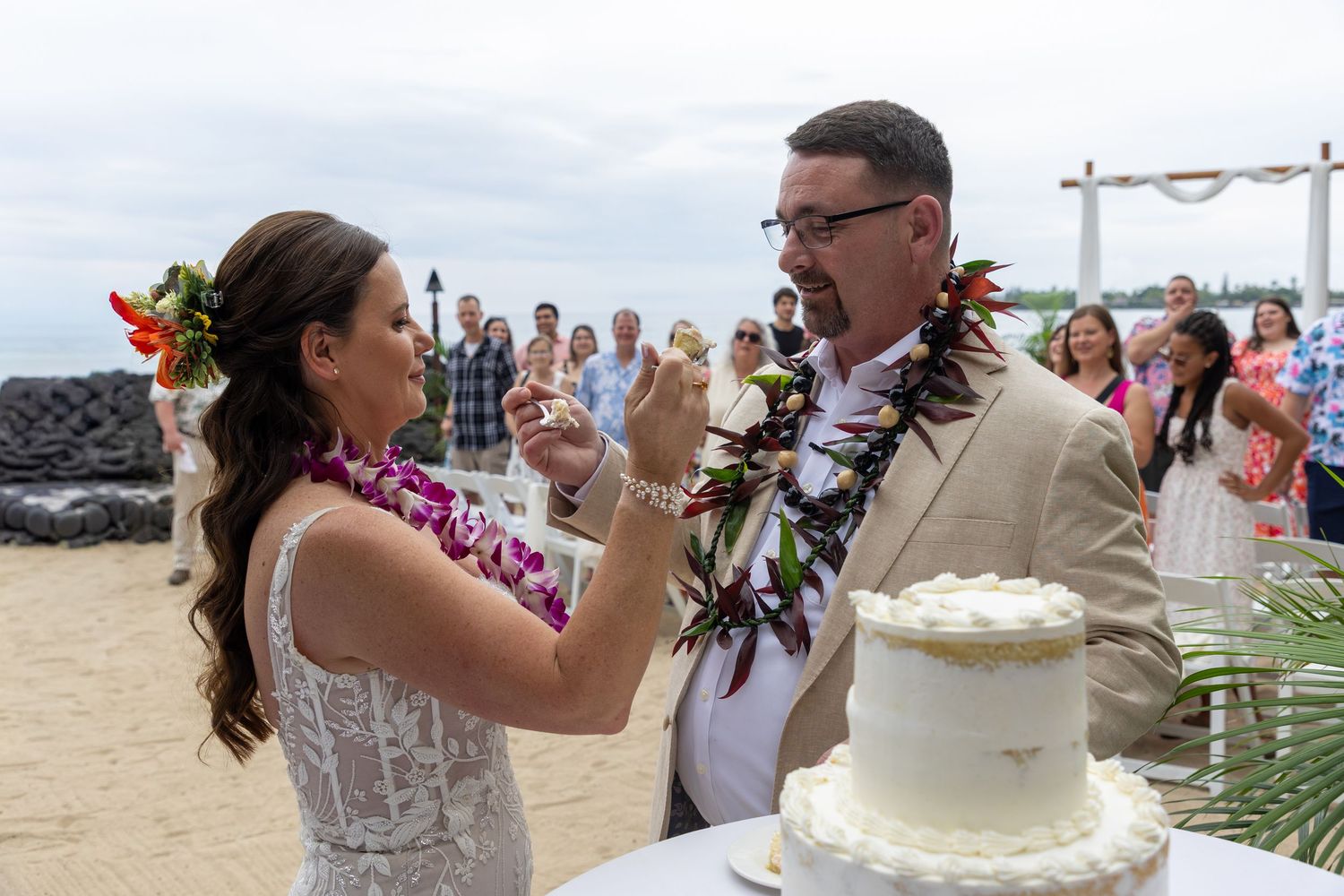 Couple cutting wedding cake on beach in Hawaii, wearing leis, with guests and ocean in background.