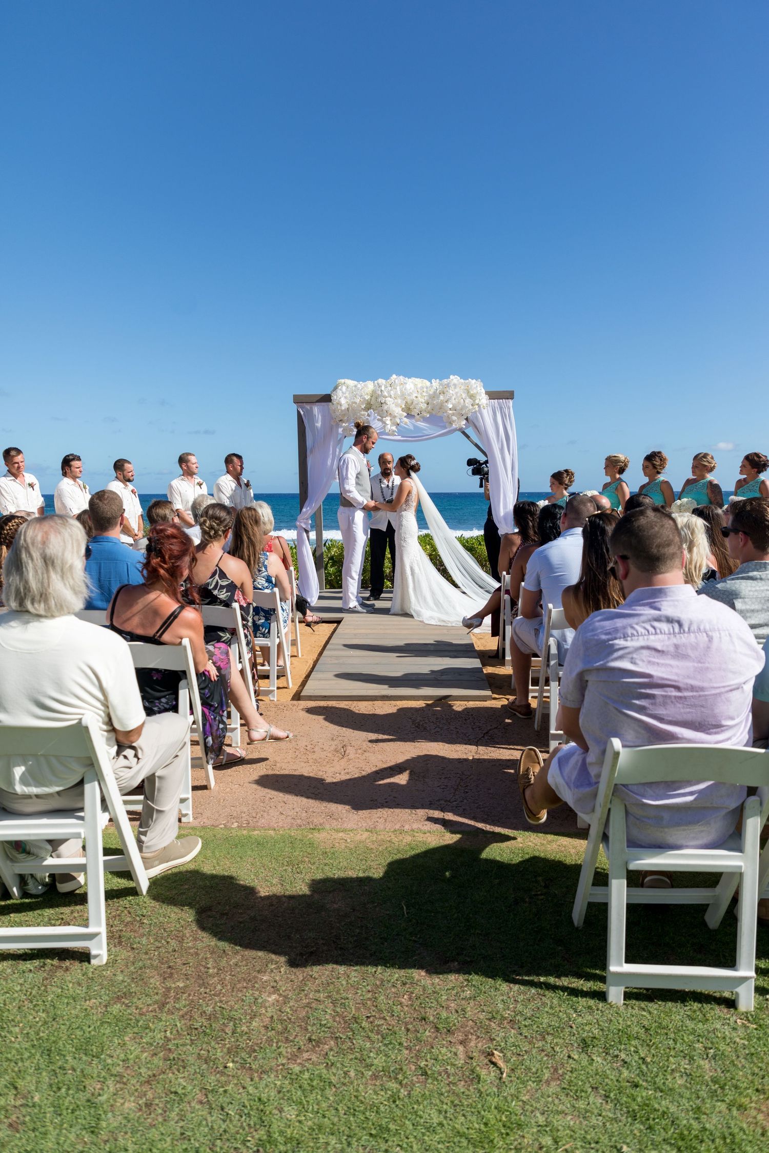 Outdoor beach wedding ceremony with guests seated in white chairs, couple at floral arch, ocean backdrop on sunny day.