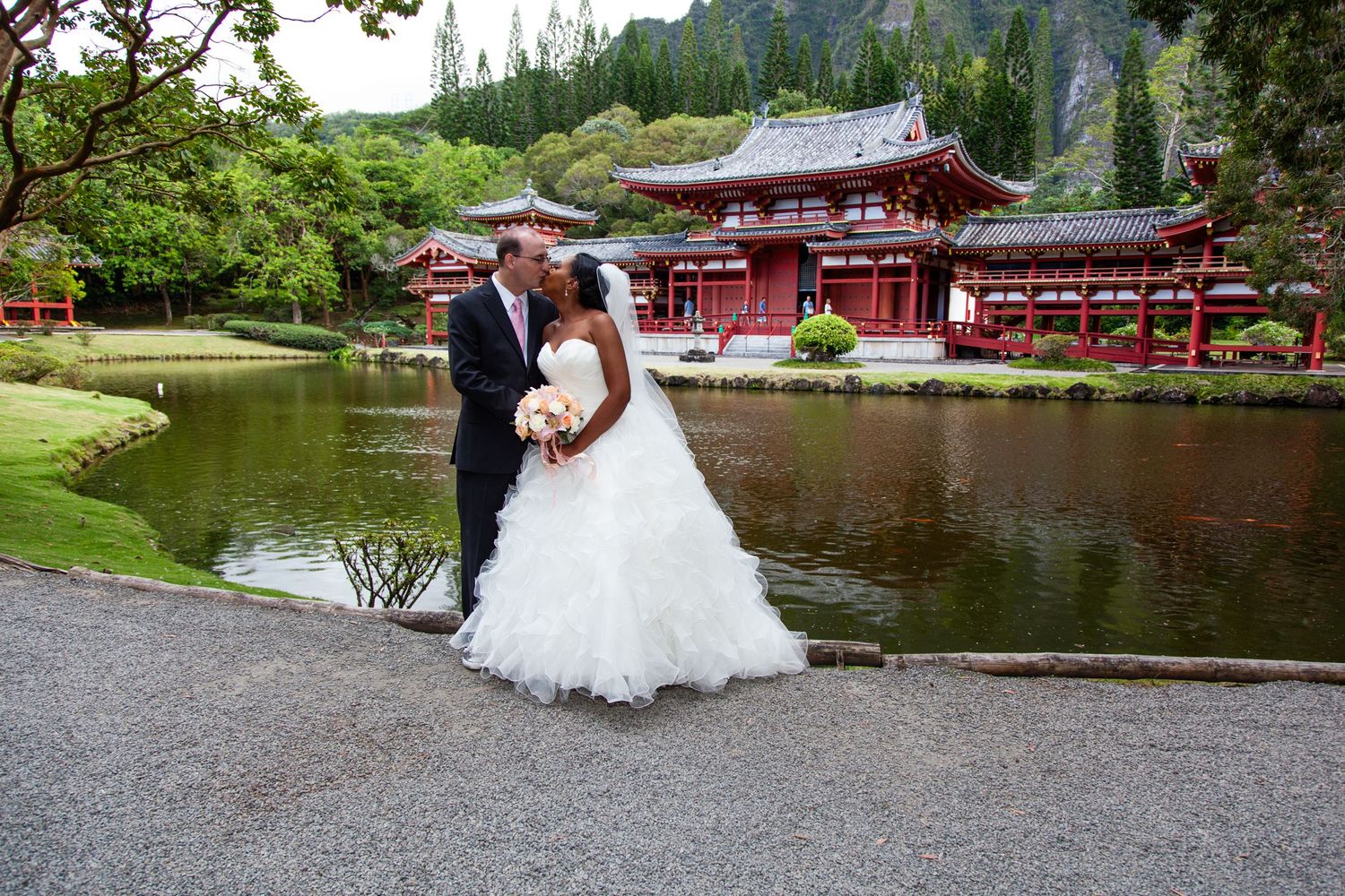 A couple in wedding attire poses near a serene pond with a traditional red Japanese temple building in the background.