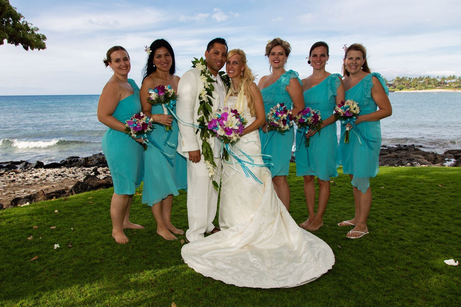 A beachside wedding party in bright turquoise dresses poses together on a grassy lawn with ocean views.