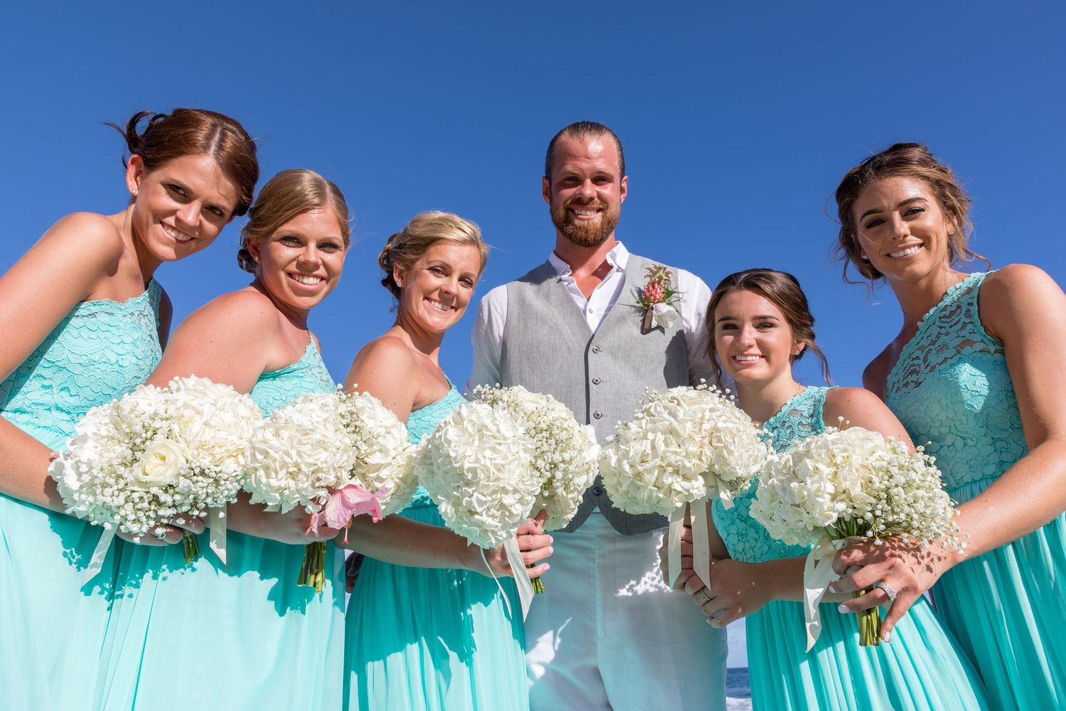 Groom in gray suit with five bridesmaids in teal dresses holding white bouquets outdoors under a clear blue sky.