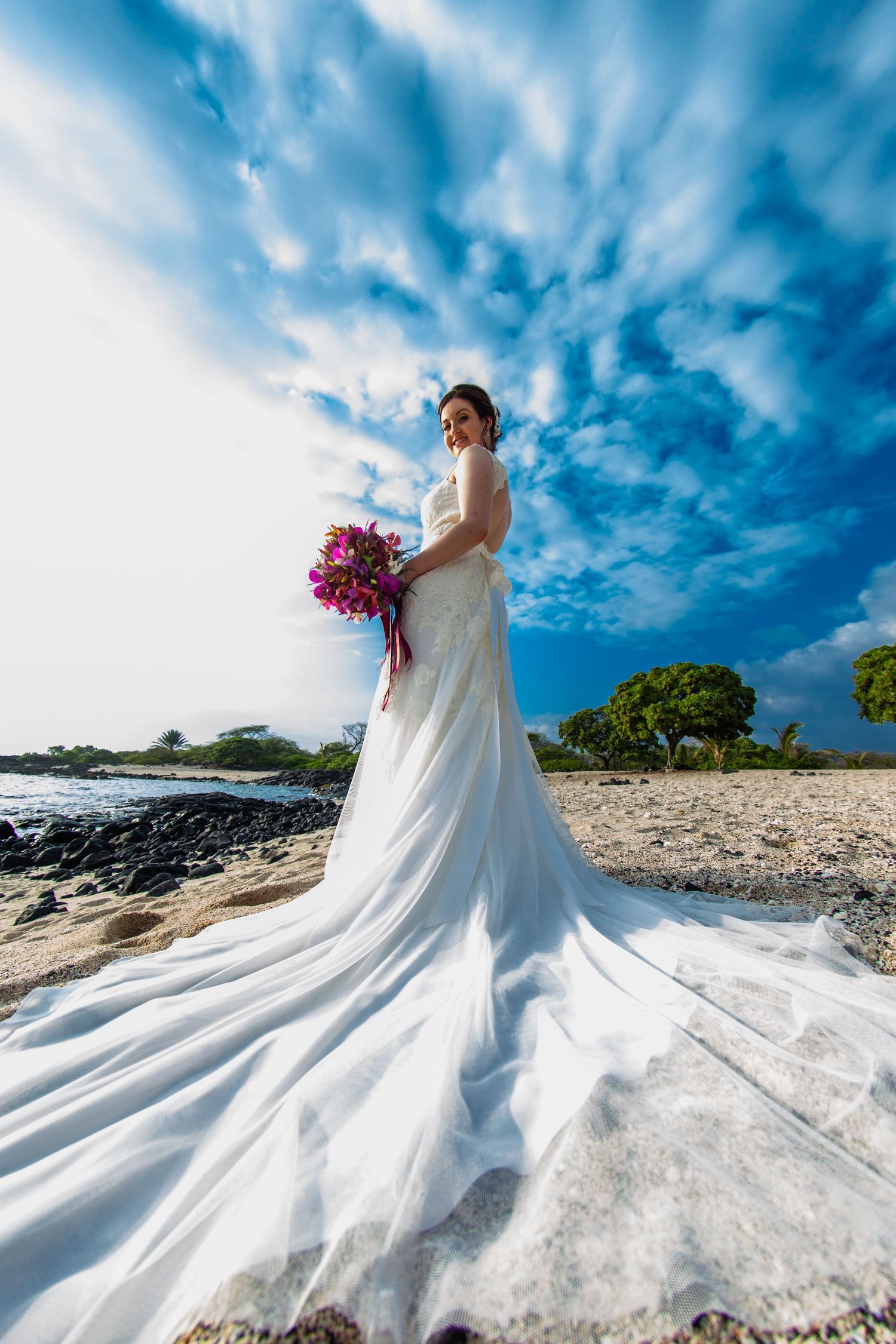 Dramatic bridal portrait on beach with flowing white dress against blue cloudy sky in Kailua Kona.