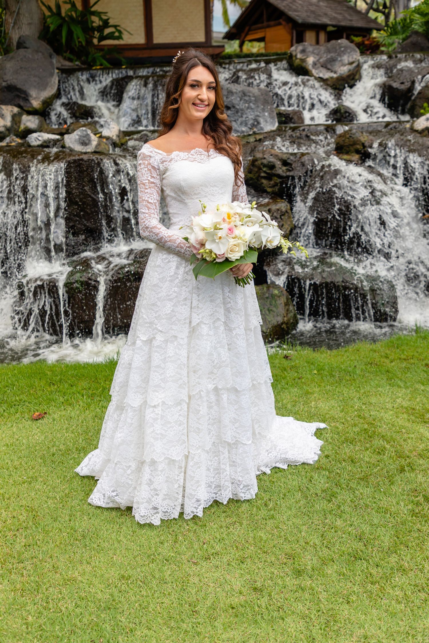 A bride in a lace wedding dress poses in front of a cascading waterfall surrounded by tropical landscaping at a hotel in Honolulu.