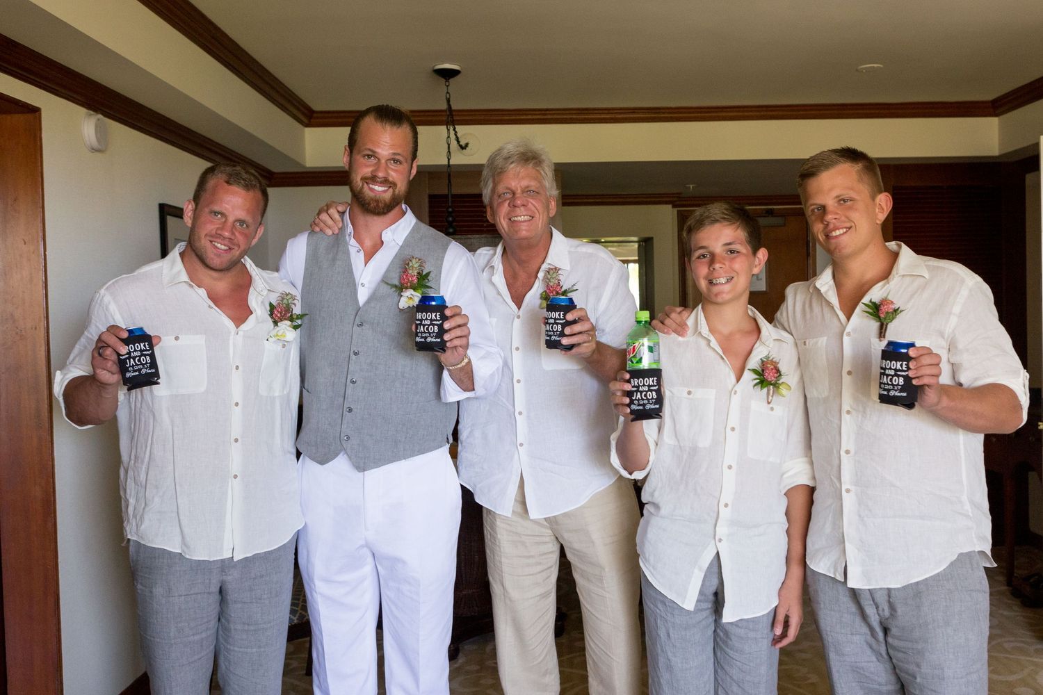 Five men in white shirts with boutonnieres smile and hold drinks at a sunny wedding celebration.