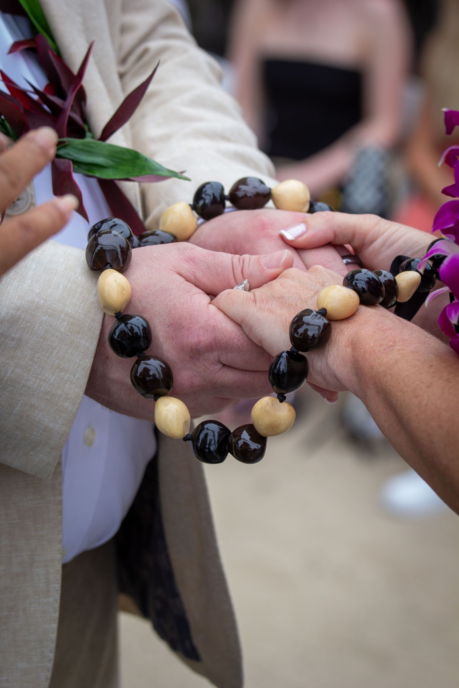 Couple holding hands with black and tan beaded bracelet during outdoor wedding ceremony.