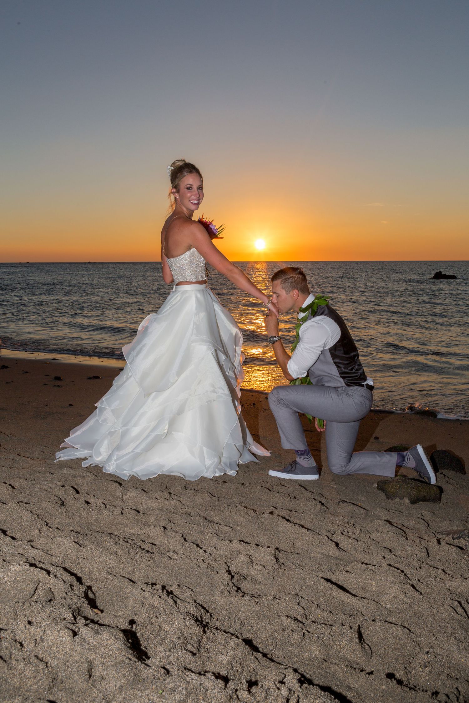 Bride and groom share romantic sunset moment on beach, groom kneeling holding bride's hand in white wedding gown.