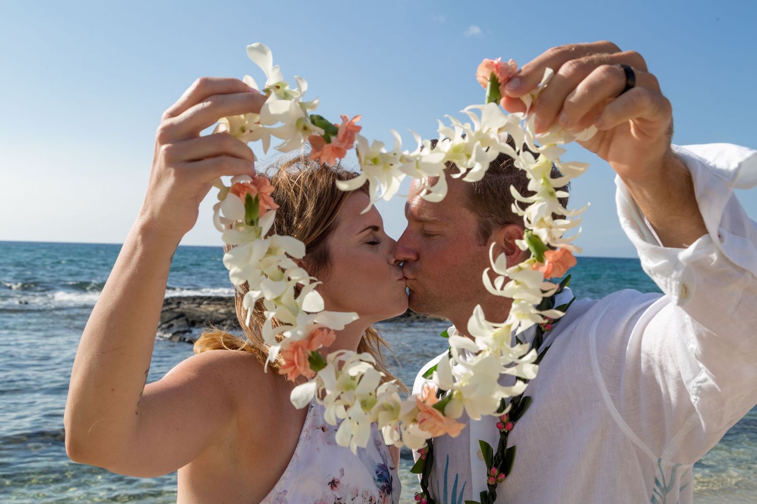 Couple kissing on beach holding white orchid lei in heart shape with ocean backdrop on sunny day.