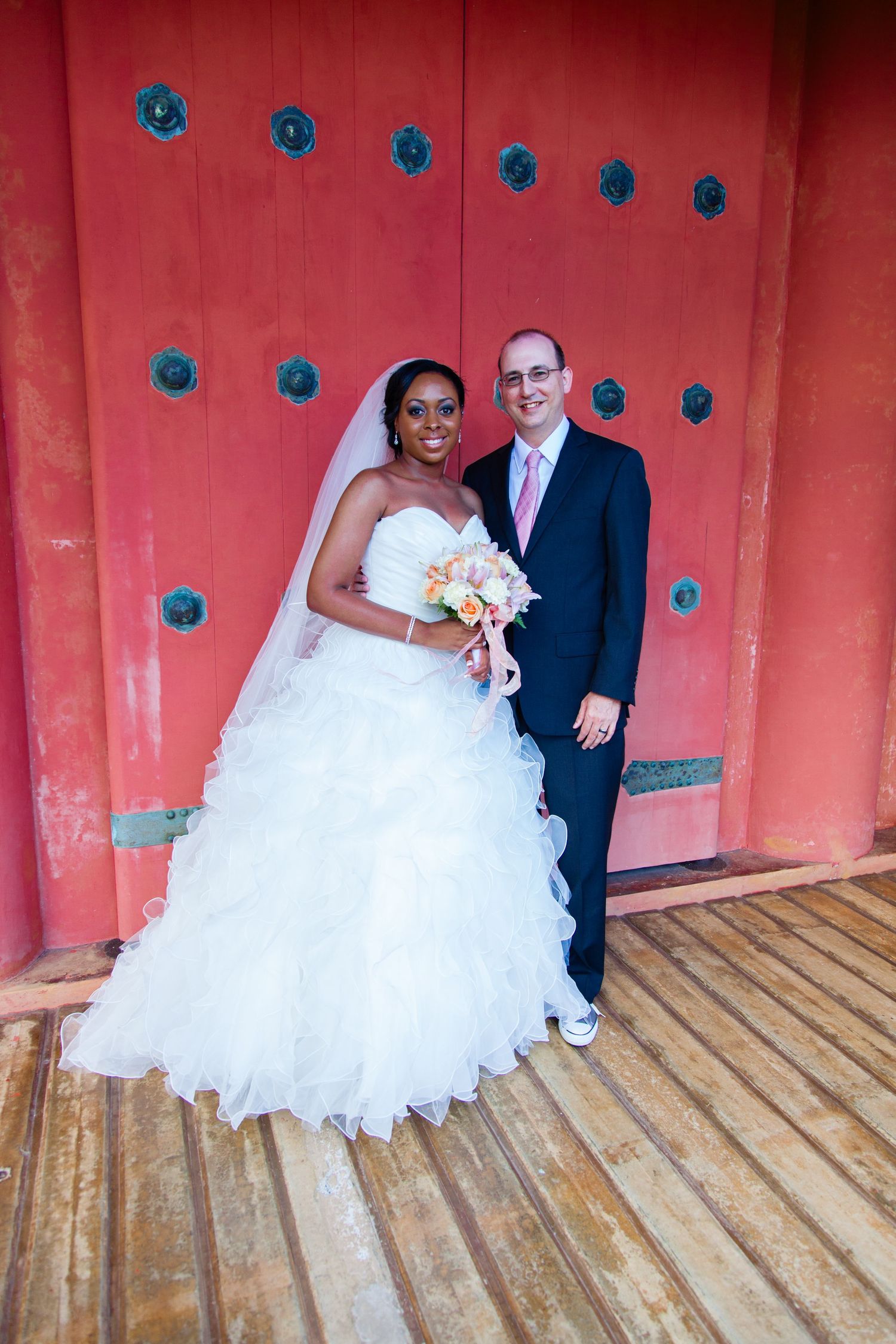 Wedding couple poses against a vibrant coral-colored wall with blue decorative accents on a wooden deck.