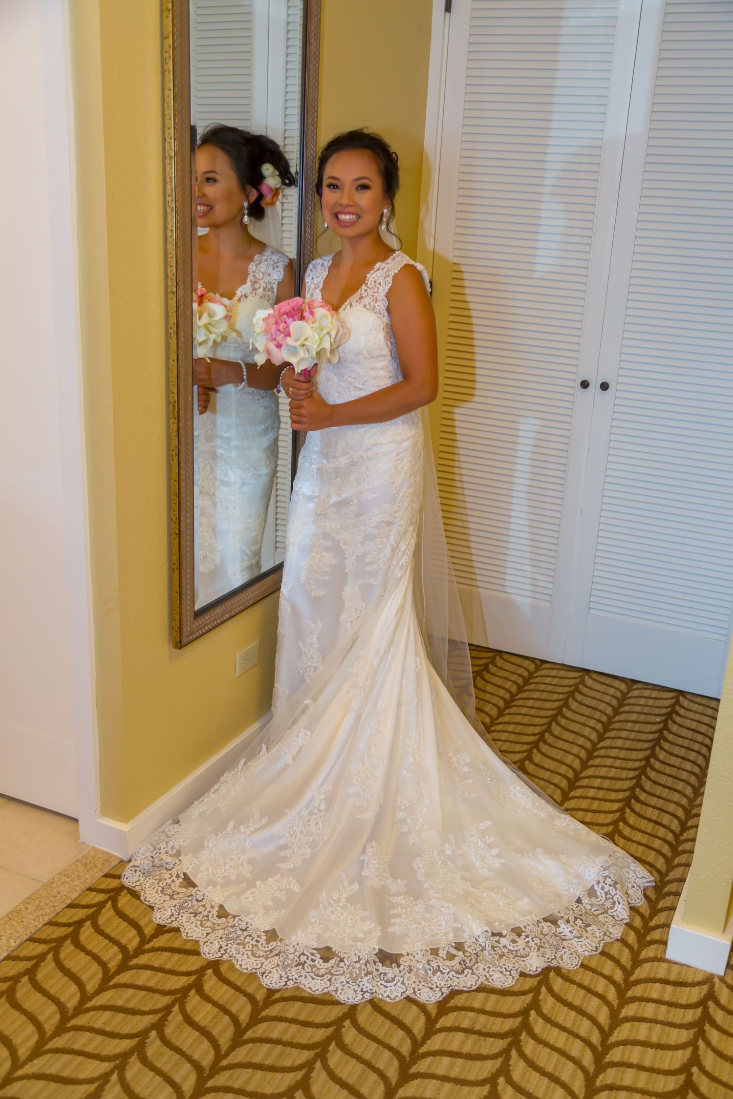 In a room of the King Kamehameha hotel in Kona, a bride stands in a white lace wedding gown with a long train in front of a mirror