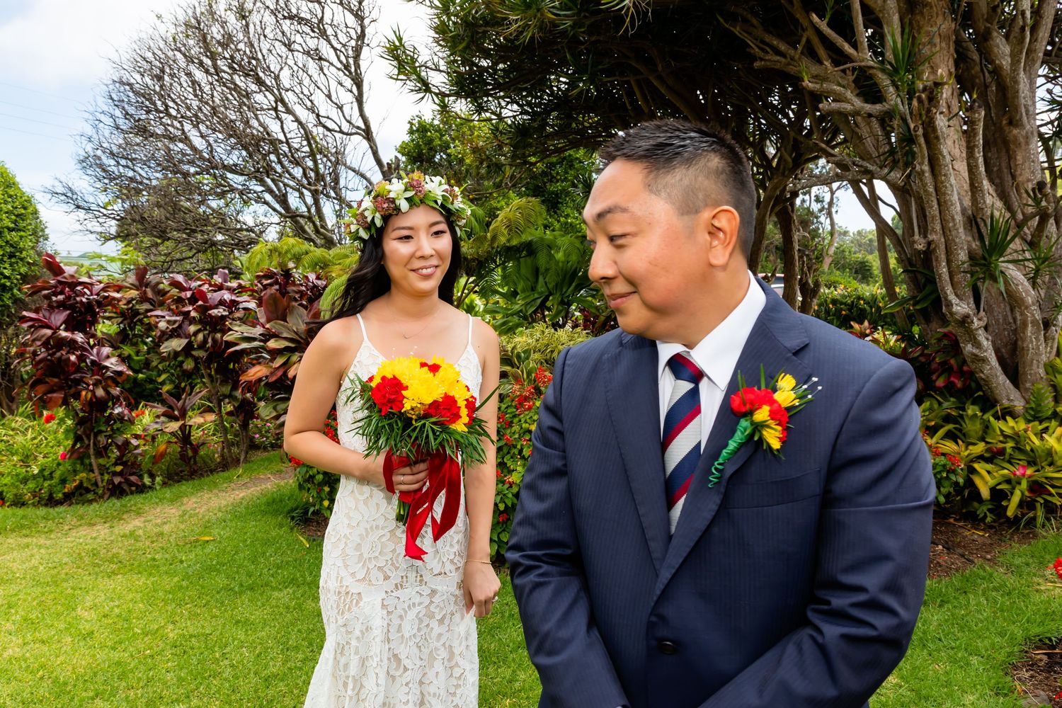 Bride in lace gown with floral crown holds tropical bouquet beside groom in navy suit with boutonniere at outdoor wedding.
