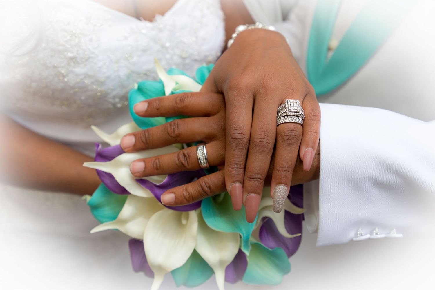 Newlywed couple displaying wedding rings on hands resting over a bridal bouquet of teal, purple, and white calla lilies.