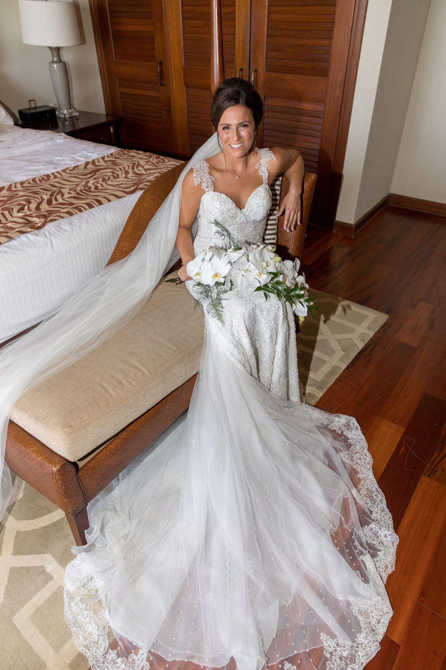 Smiling bride in a lace mermaid wedding gown holding white bouquet seated in elegant bedroom.