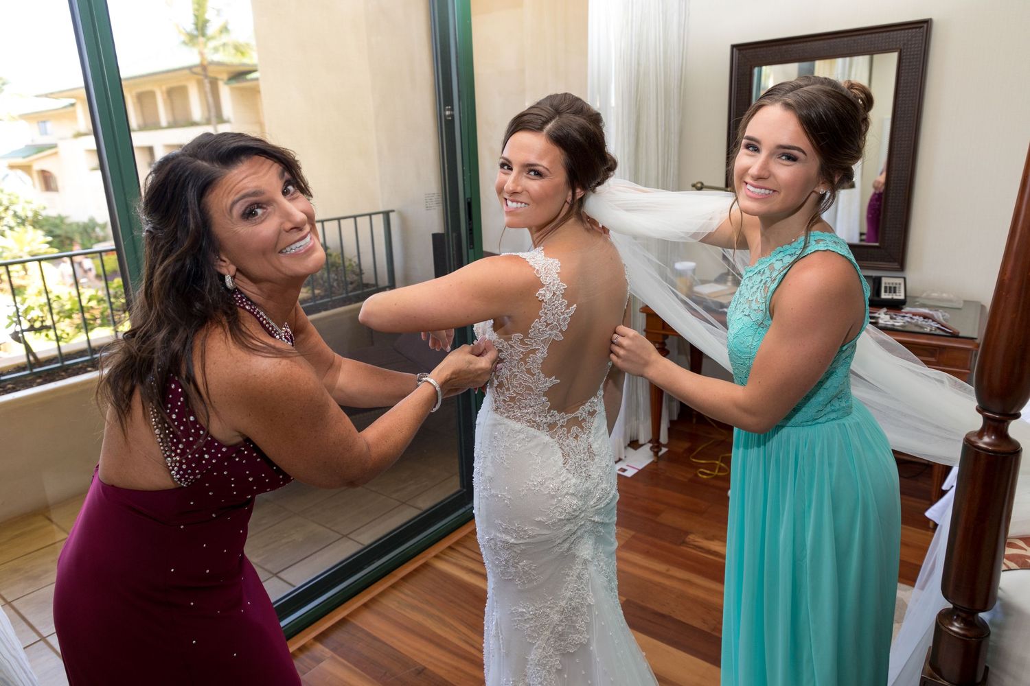 Two bridesmaids help a bride in a lace wedding gown with her dress before the wedding ceremony.