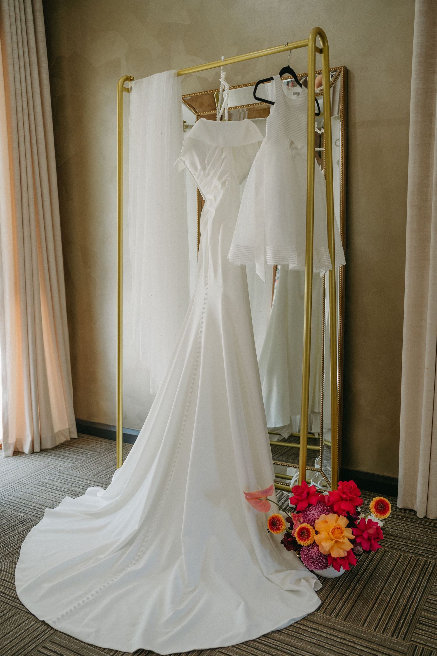 White wedding dress with long train displayed on gold rack with vibrant red and orange floral bouquet accent.