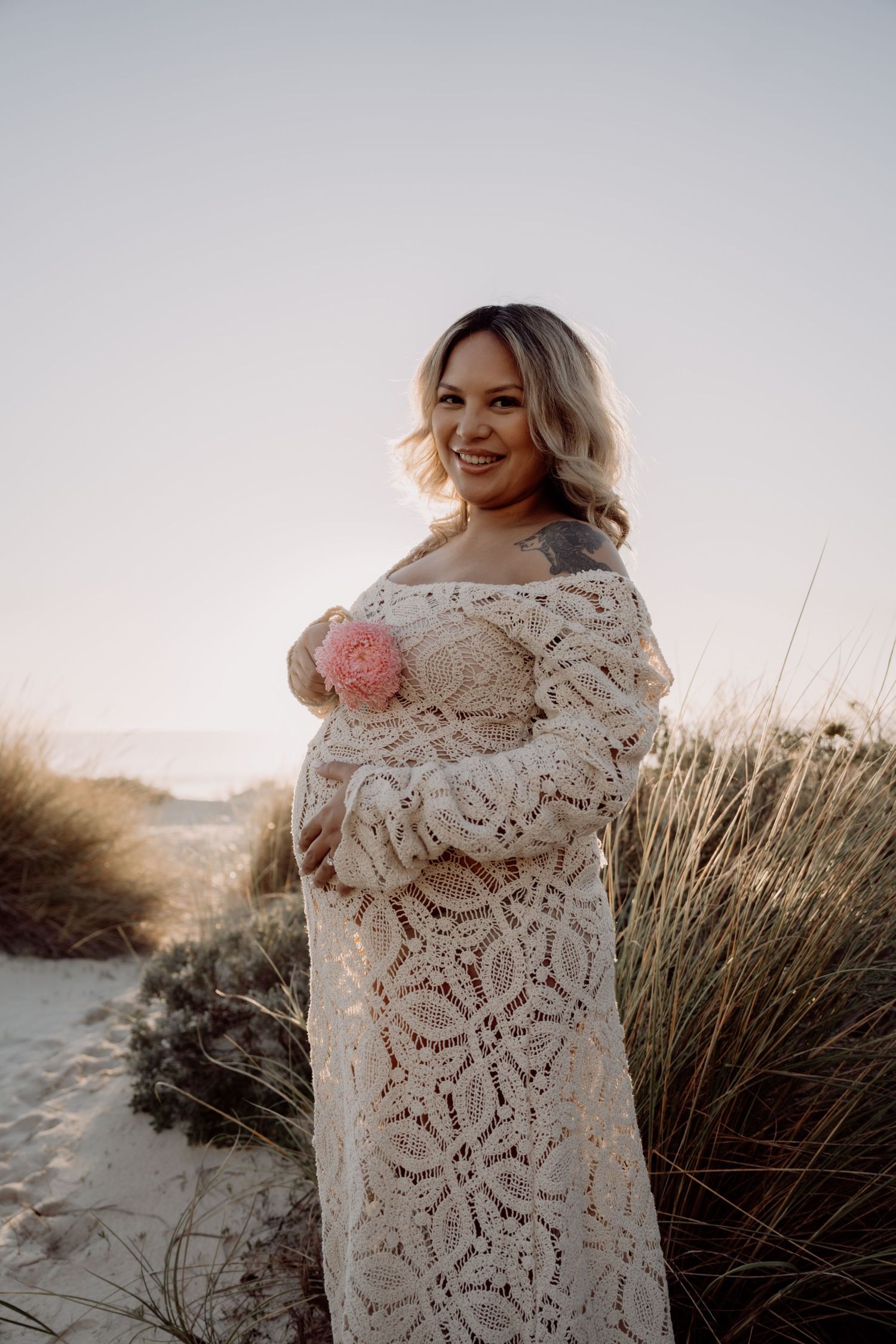 A person wearing a delicate white lace shawl poses against coastal dunes and grass at sunset.