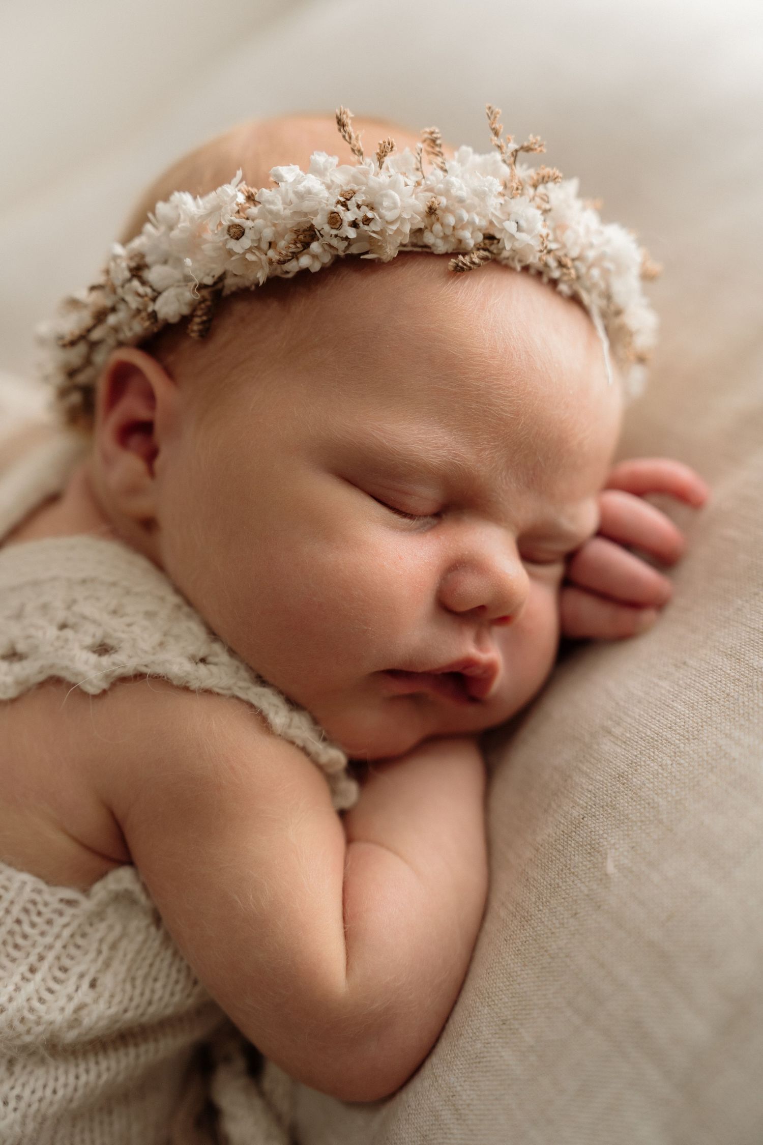 Sleeping newborn wearing delicate white floral crown and lace outfit rests peacefully on beige fabric.