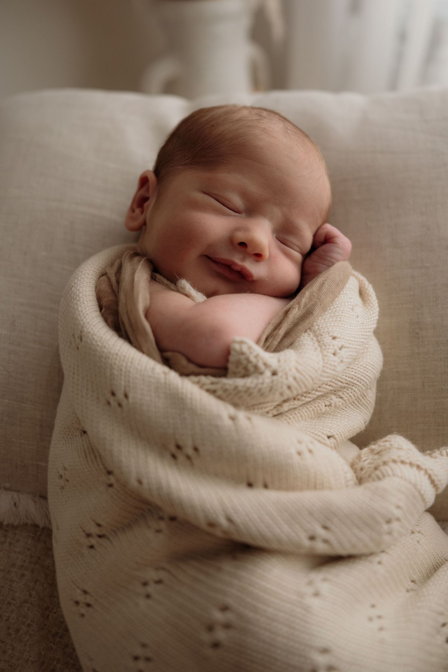 A sleeping newborn baby swaddled snugly in a soft cream-colored knit blanket in a peaceful studio setting.