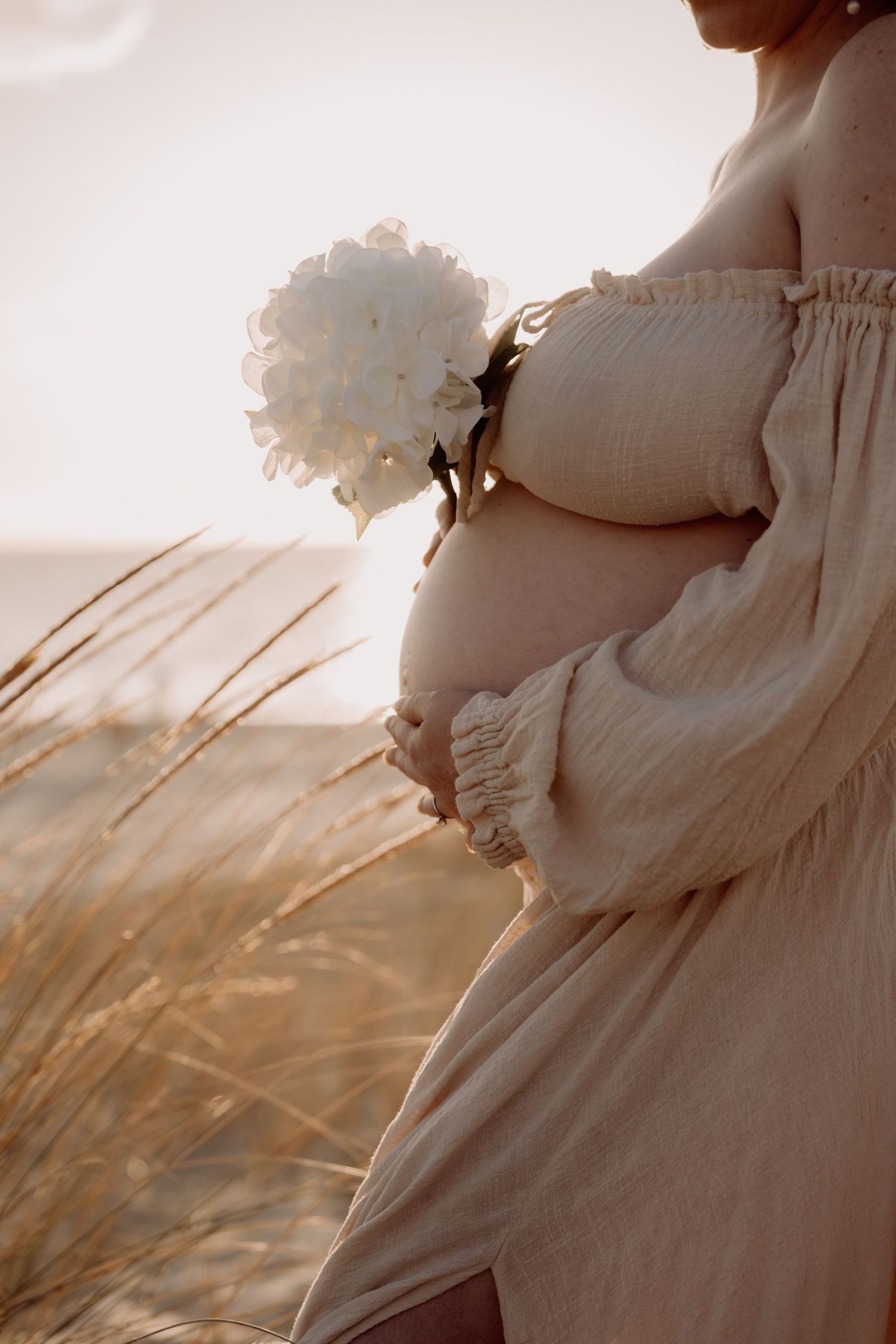 A pregnant woman in a white dress holds white flowers while standing in tall grass against golden sunlight.