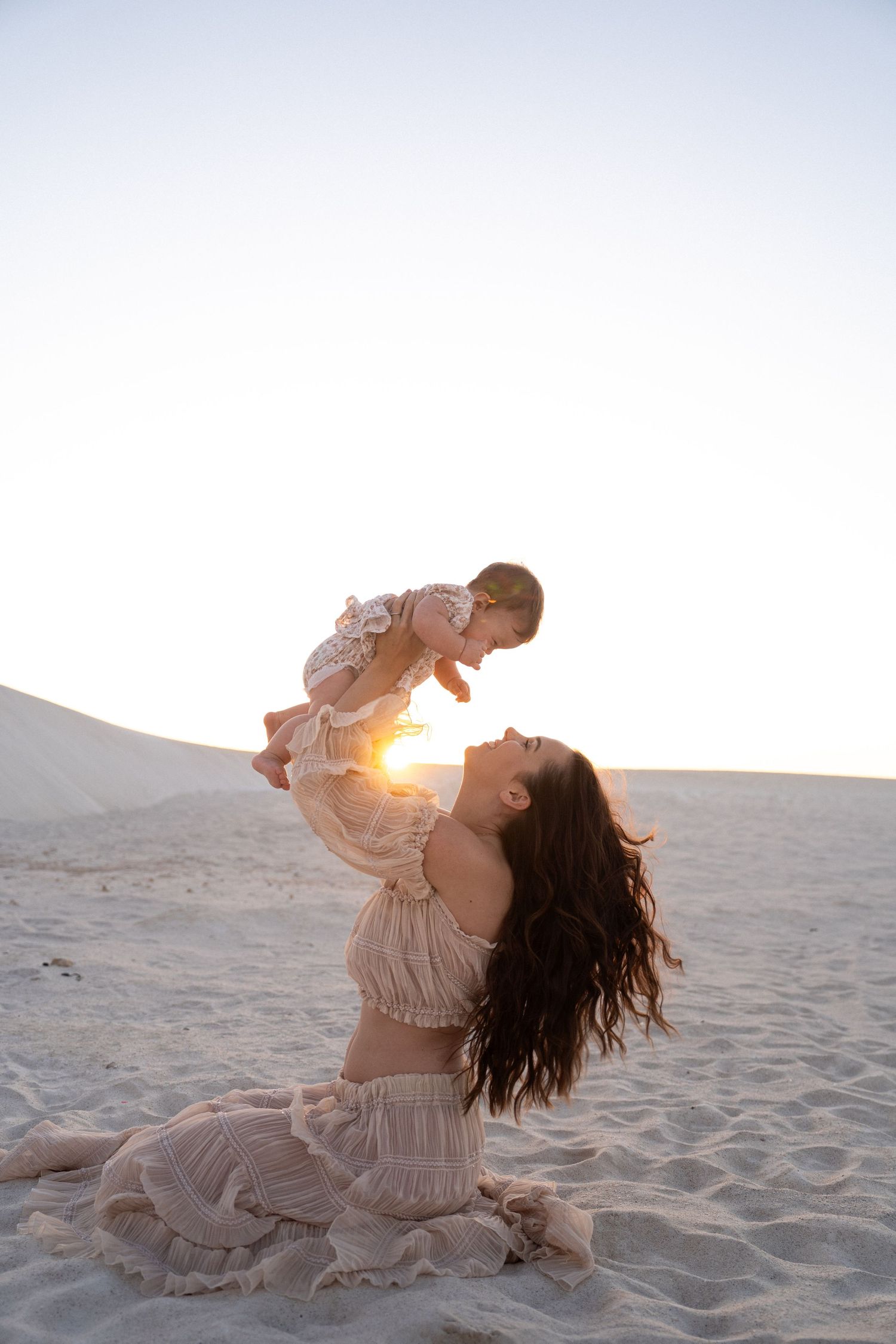A woman in a flowy cream dress lifts up a baby against a sunset backdrop on sandy desert dunes.