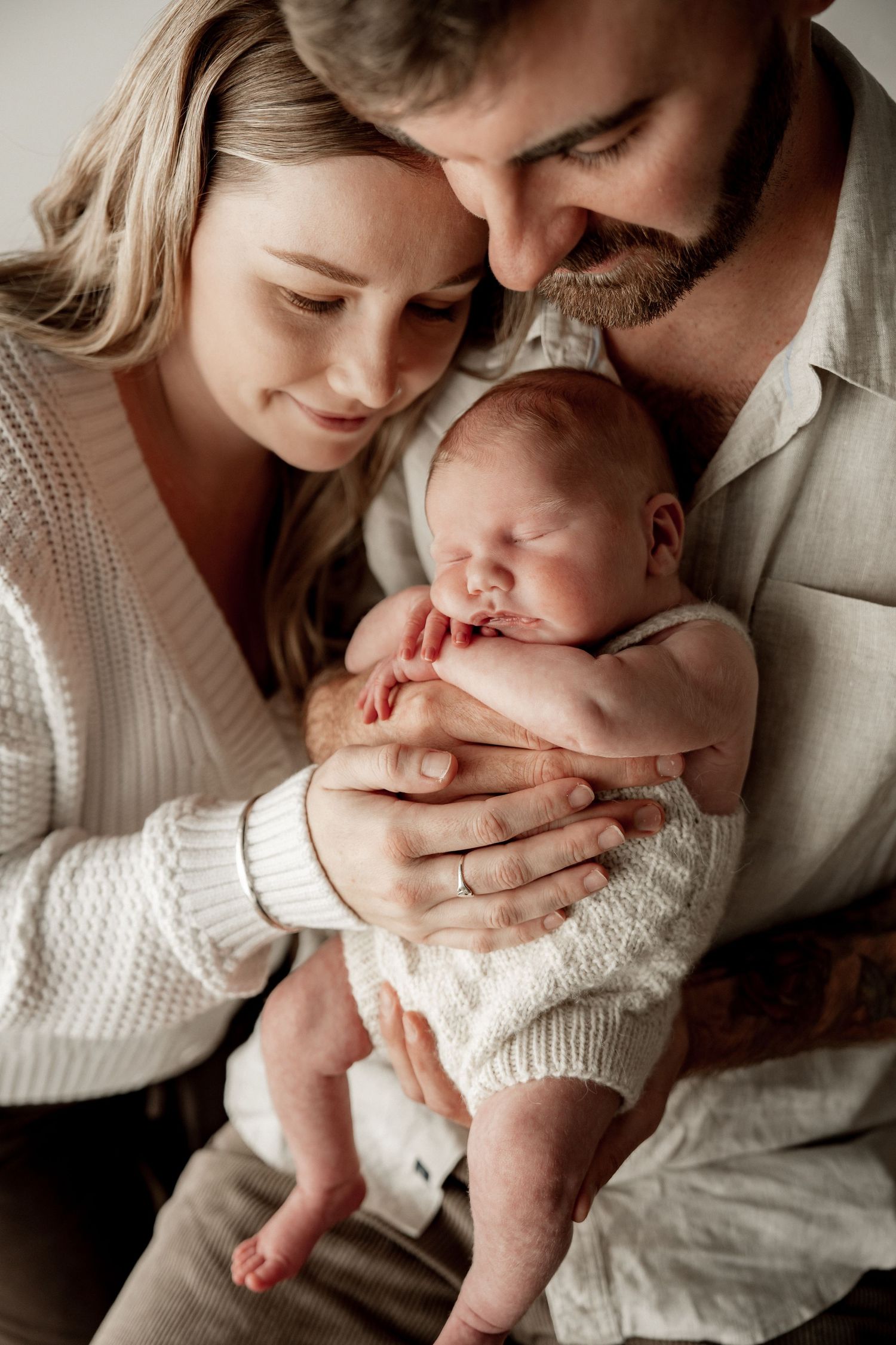 Young parents lovingly cradle their sleeping newborn baby, sharing a tender family moment in soft neutral tones.