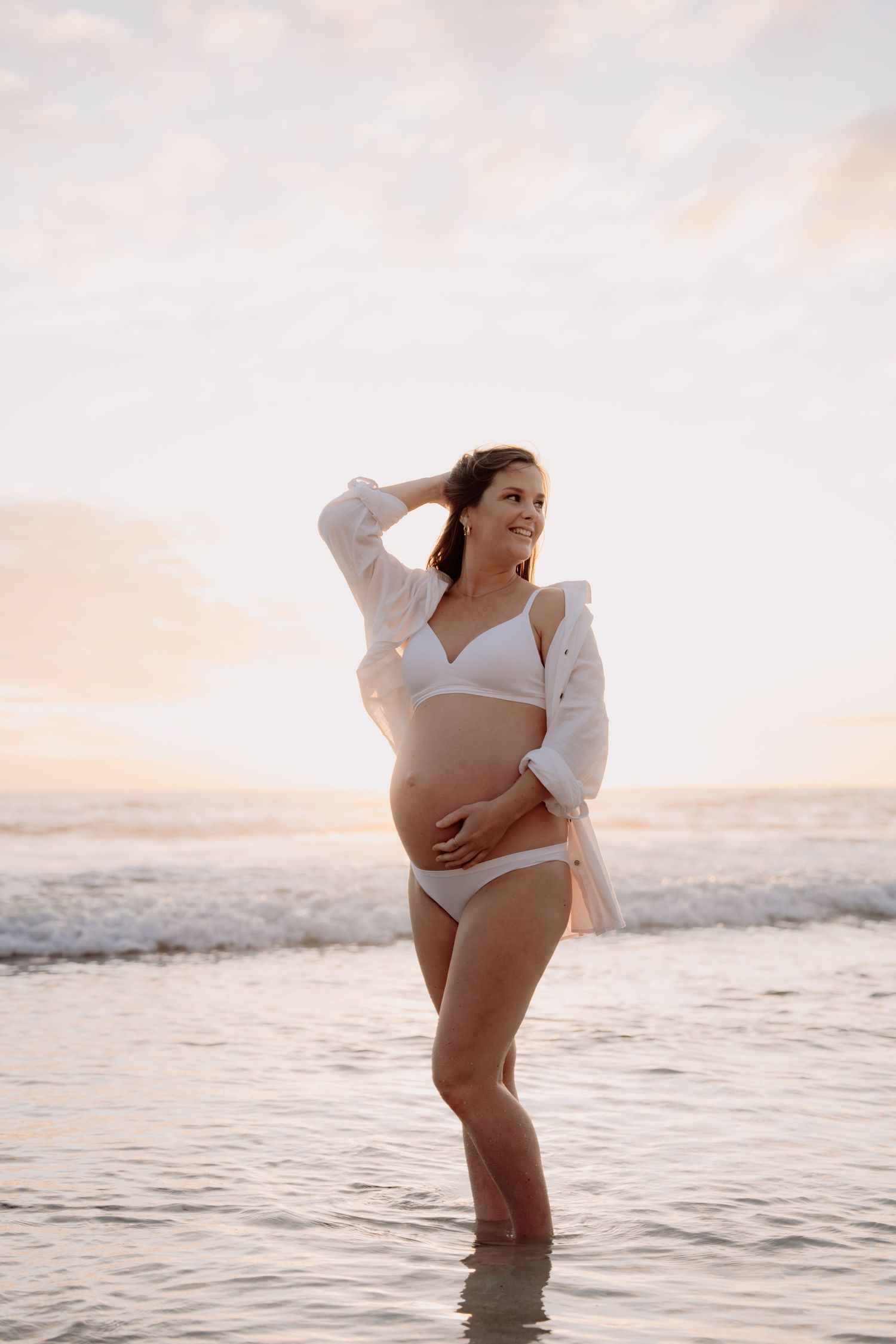 Pregnant woman in white bikini and open shirt standing in ocean waves at sunset, cradling her baby bump.