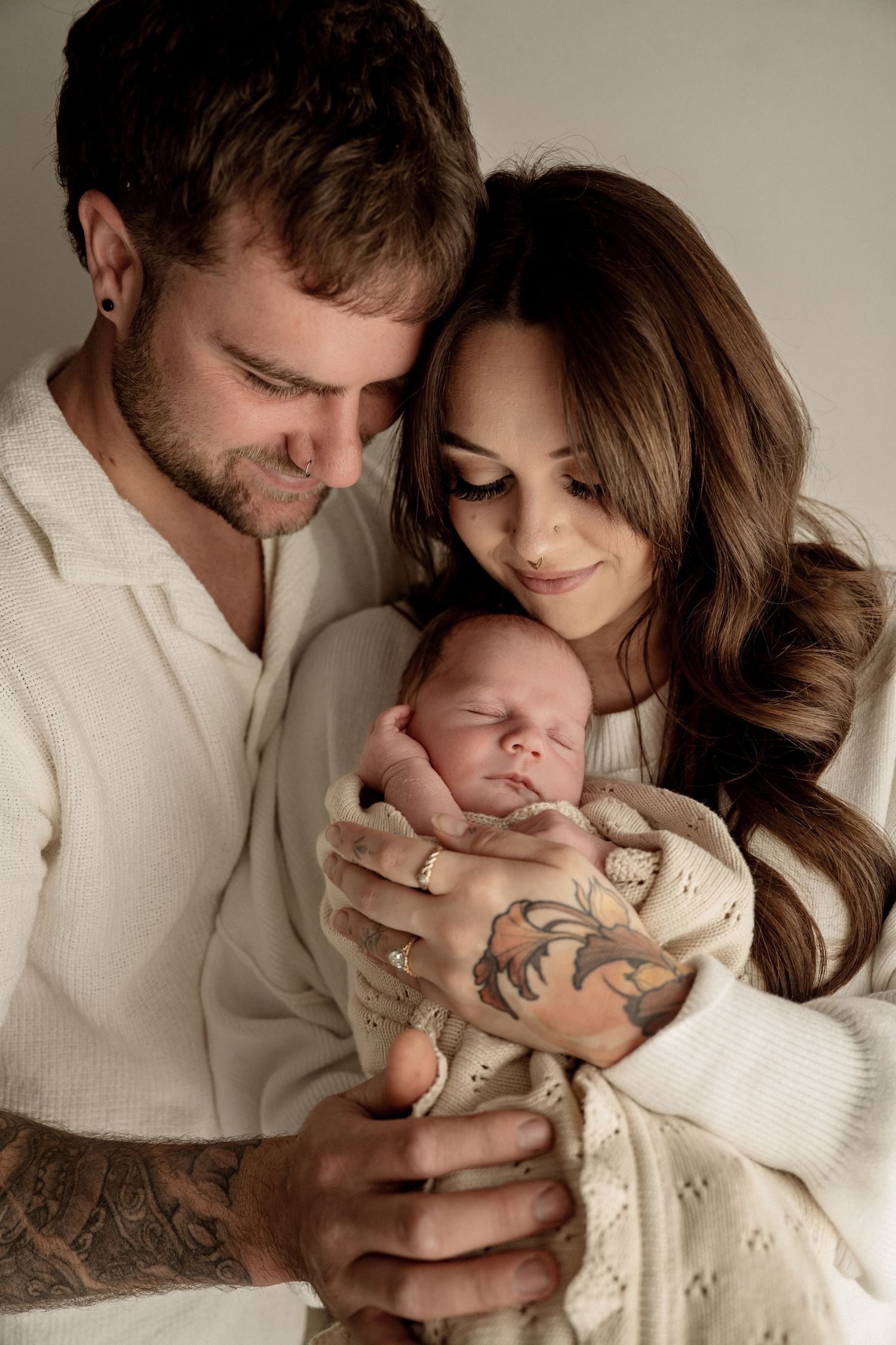 A couple in white sweaters lovingly hold their sleeping newborn baby while showcasing tattoos in a warm toned portrait.