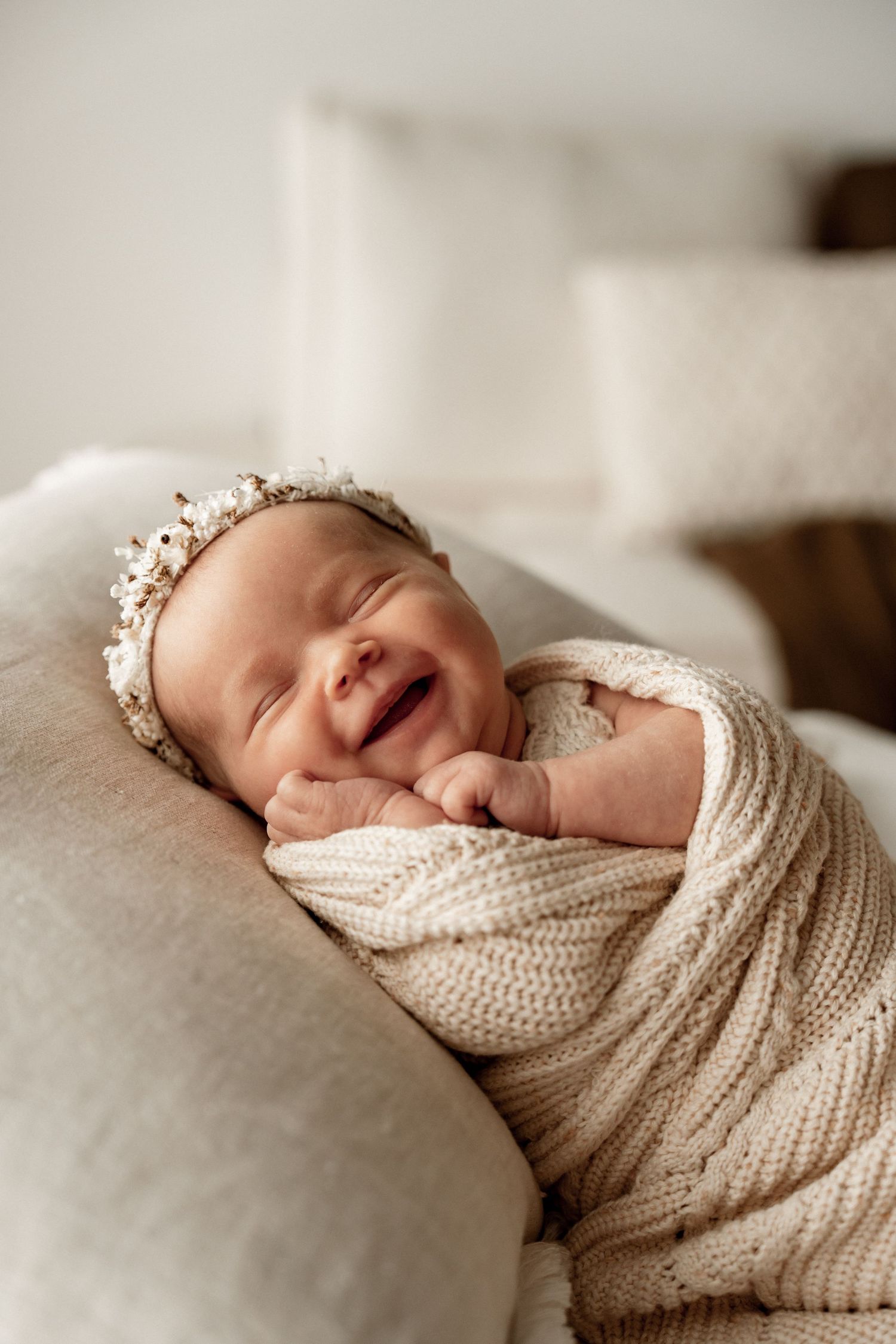 A newborn baby wrapped in a cozy cream knit blanket smiles while sleeping peacefully on soft bedding.