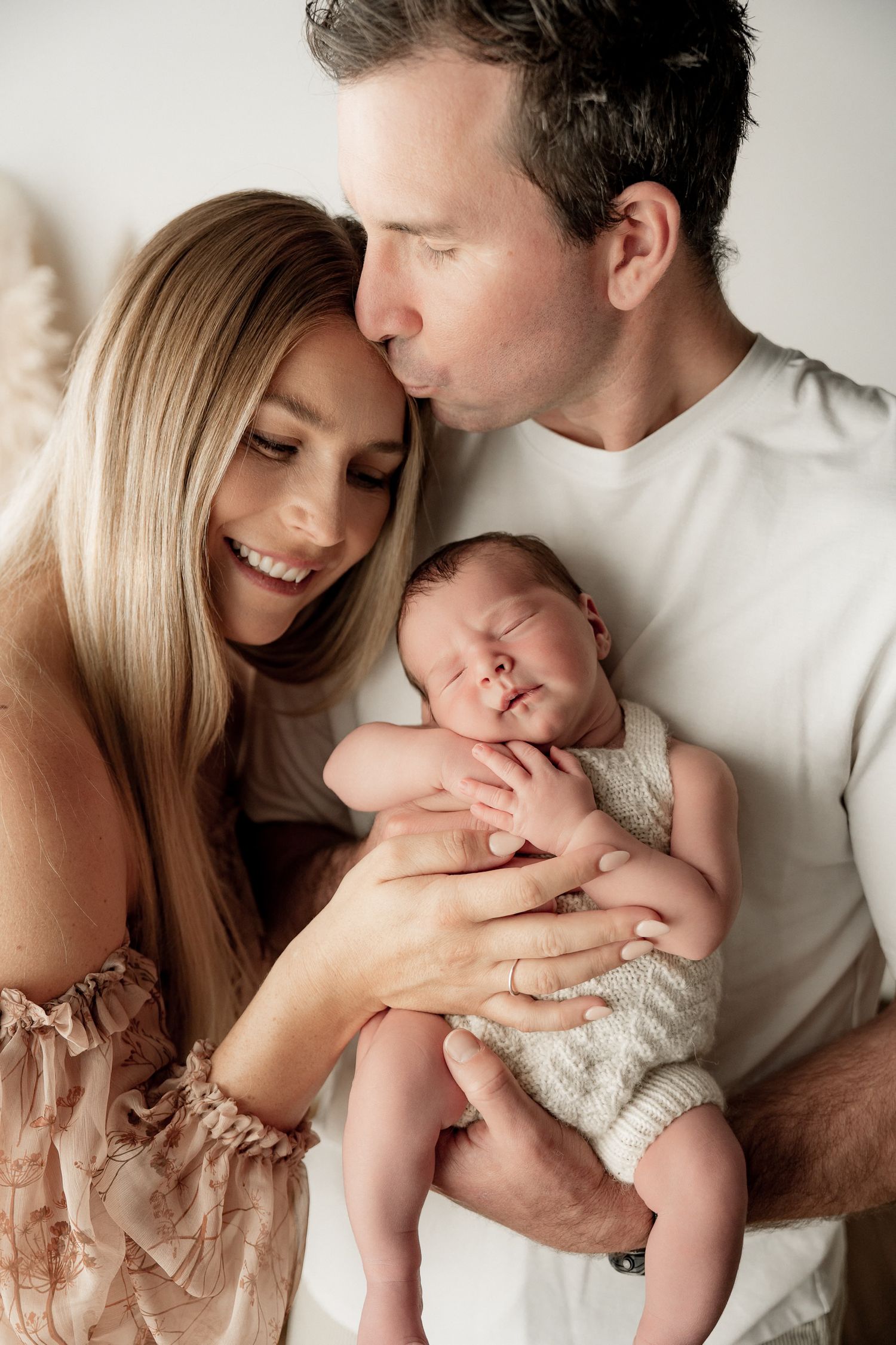 A couple smiles lovingly at their sleeping newborn baby, with the father kissing the infant's head in a warm studio portrait.