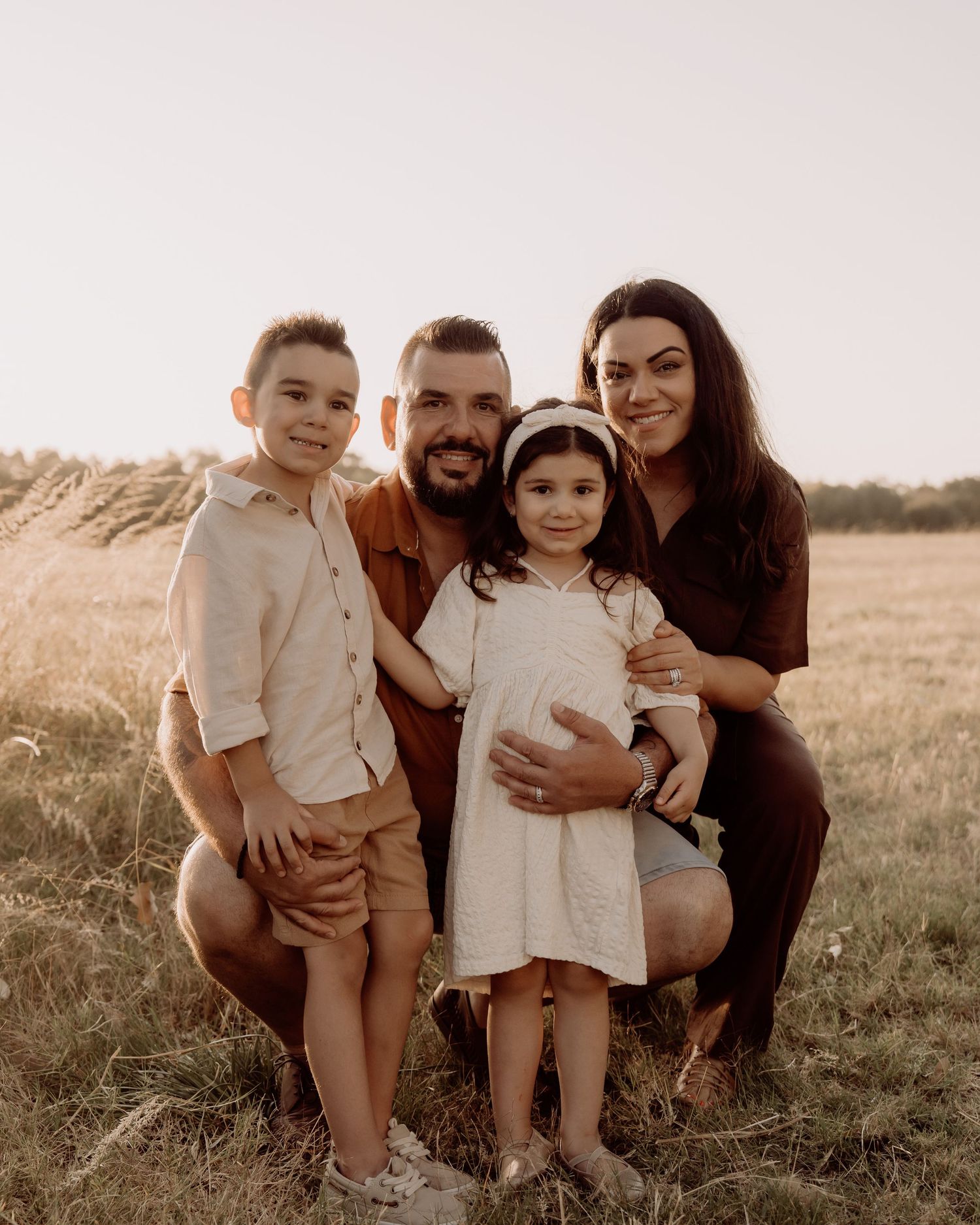 A family poses together in a sunny field during golden hour, wearing neutral colored clothing.