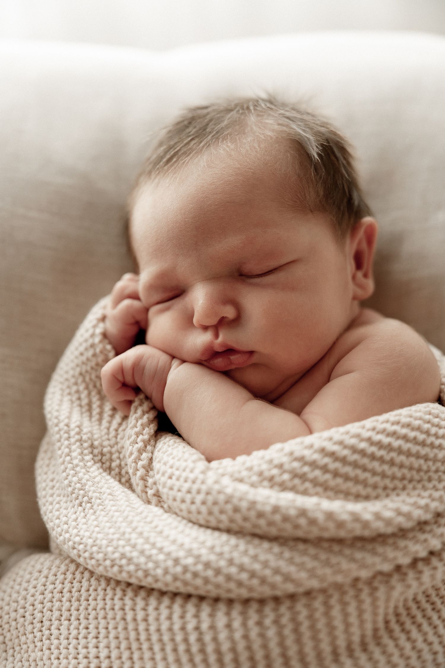Peaceful newborn baby sleeping wrapped in a soft beige knit blanket with tiny hands tucked under chin.