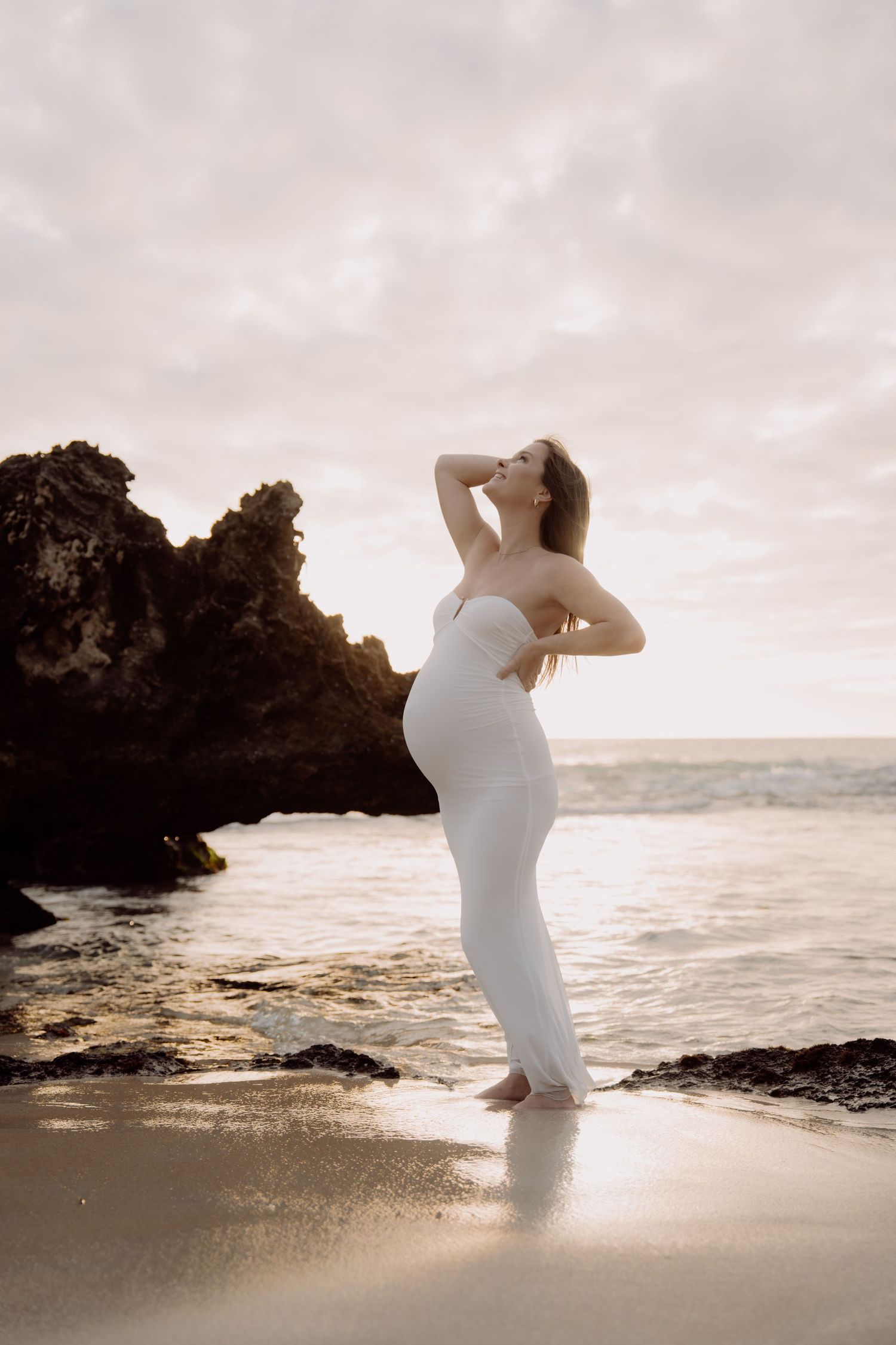 Pregnant woman in white dress posing on beach at sunset with rocky shore and ocean in background.