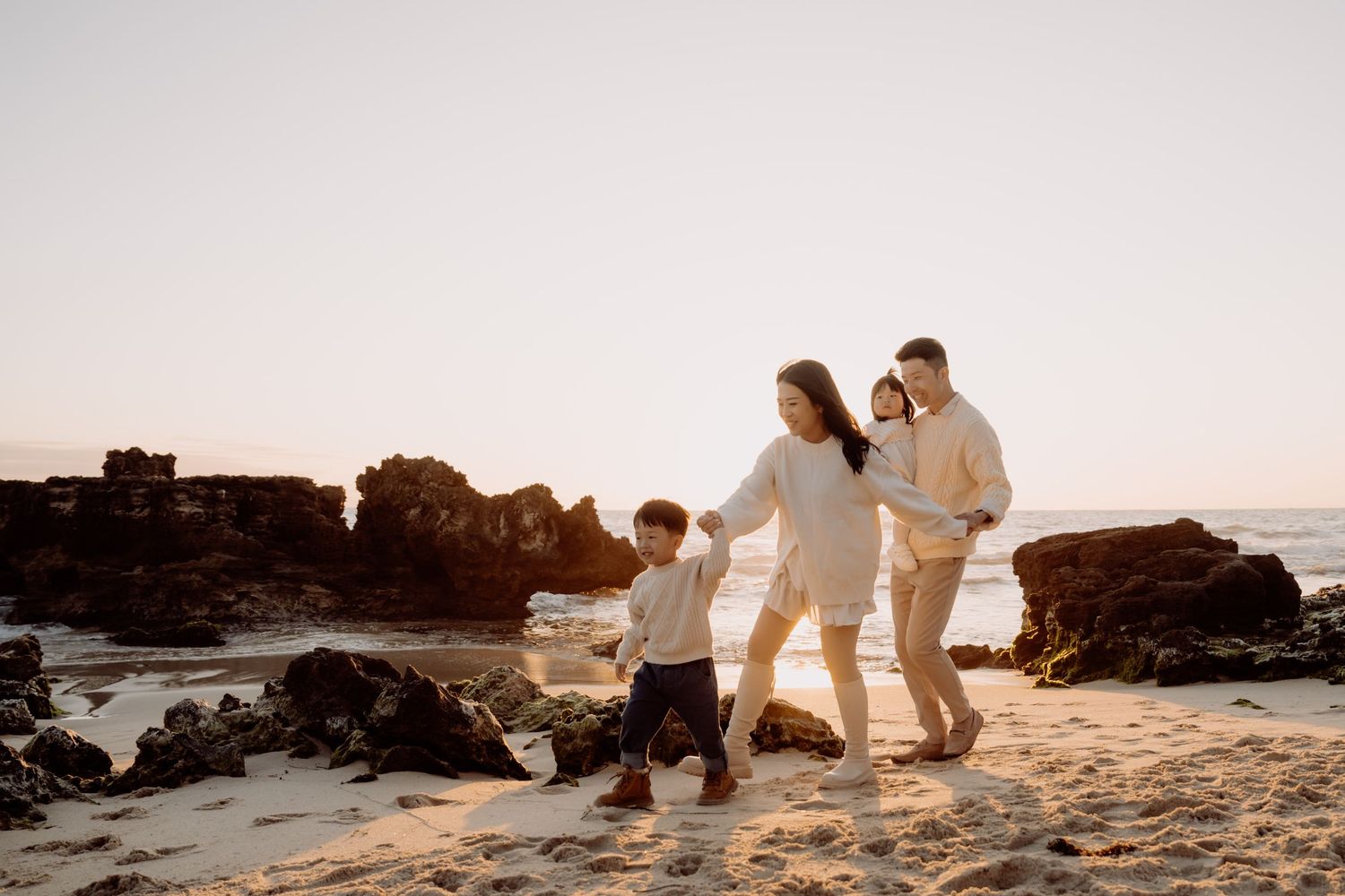 A silhouetted group walks together along a rocky beach at sunset holding hands.