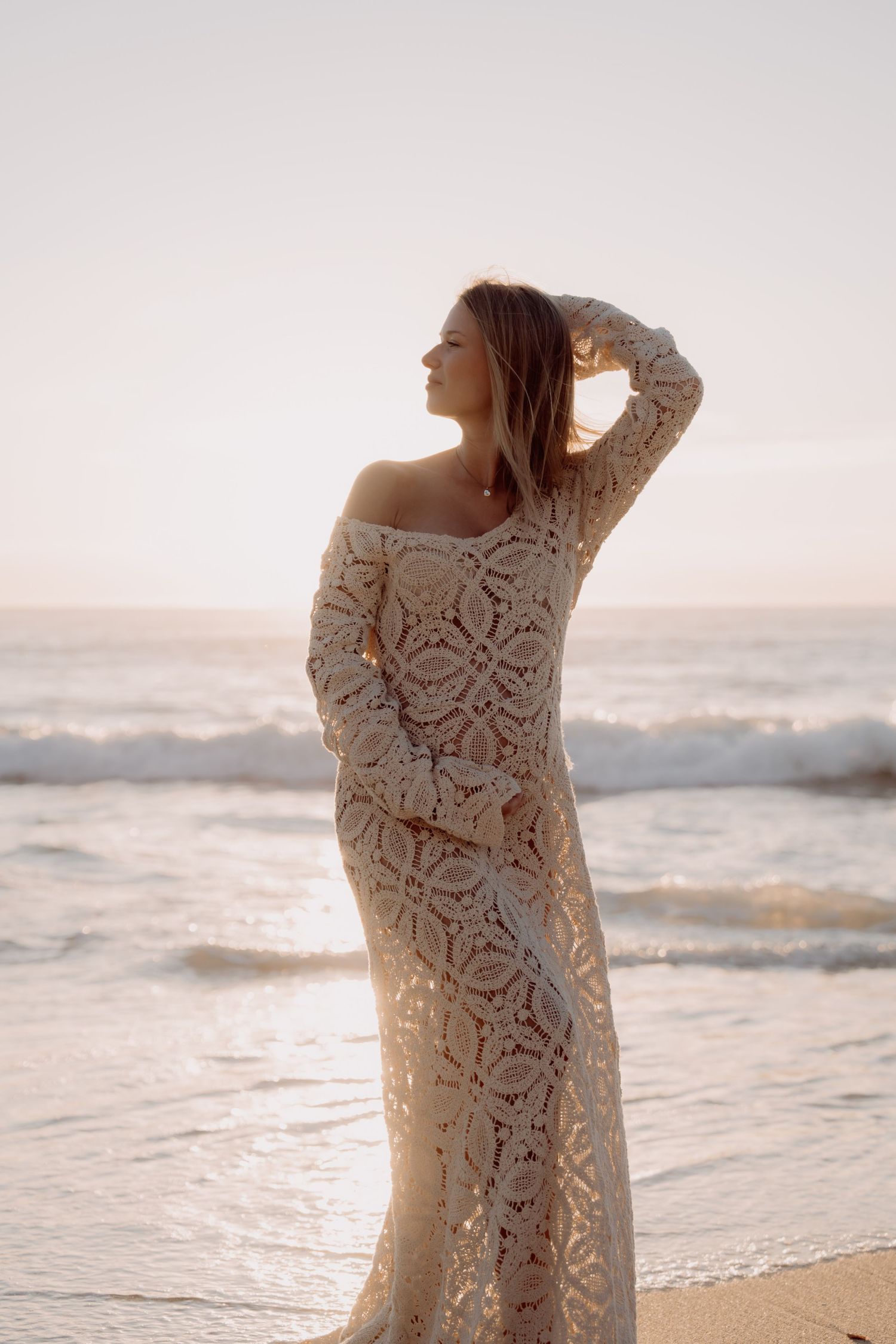 A person in a delicate lace dress poses gracefully on a beach at sunset, creating a romantic silhouette against the waves.