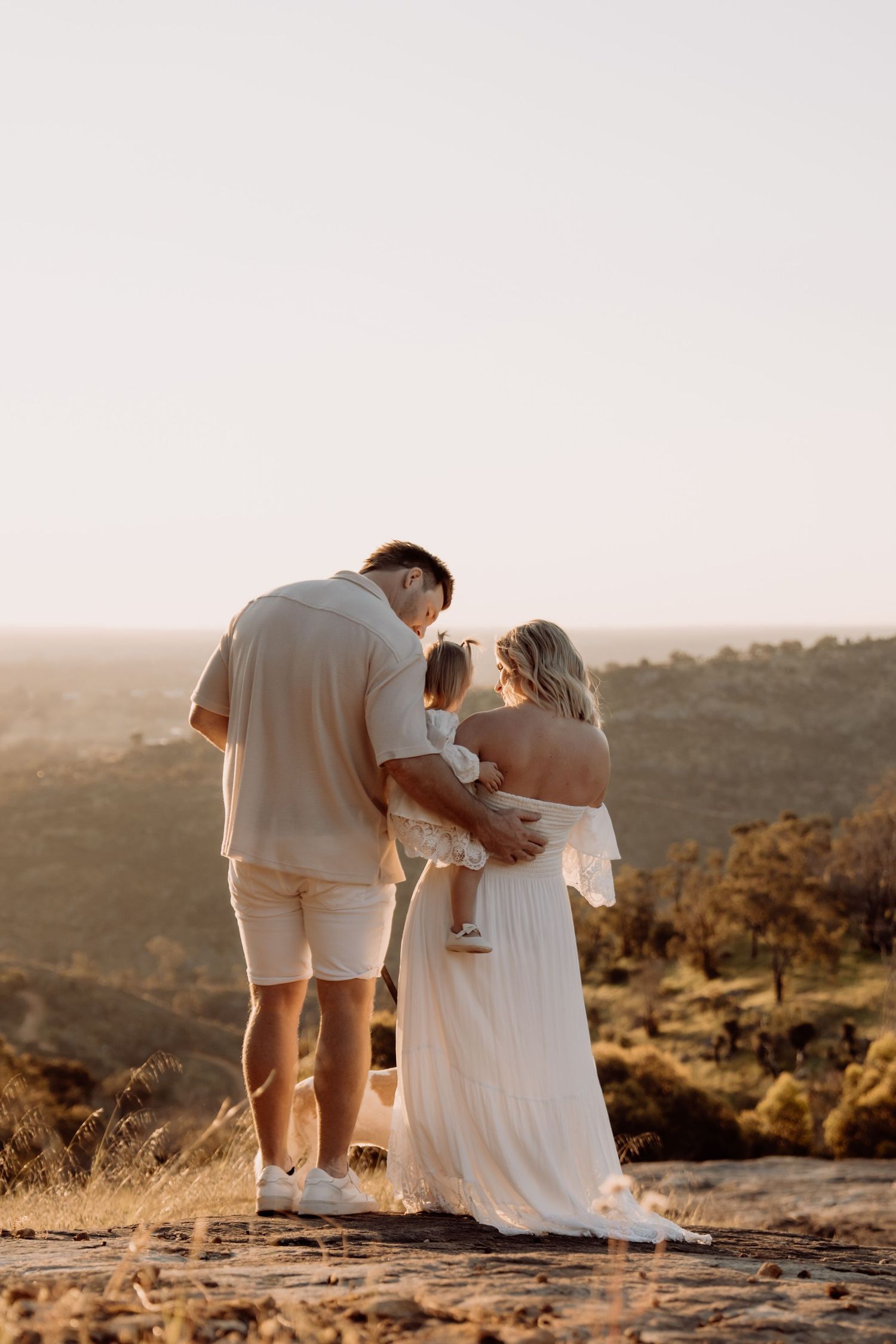 A couple in white and beige outfits share an intimate moment at sunset on a rocky hilltop.