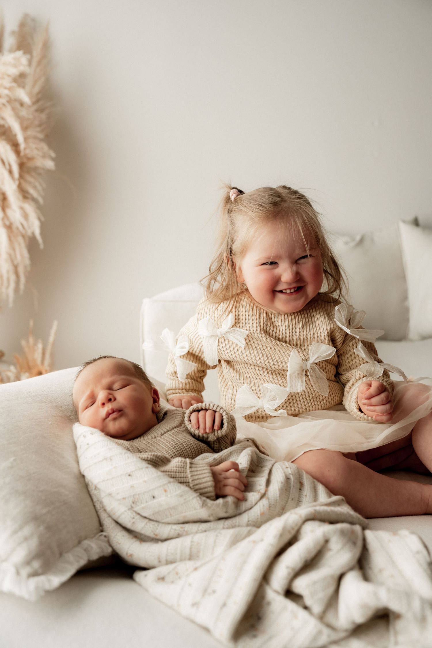 Smiling toddler girl in striped sweater sits beside sleeping newborn baby wrapped in a soft blanket on a white bed.