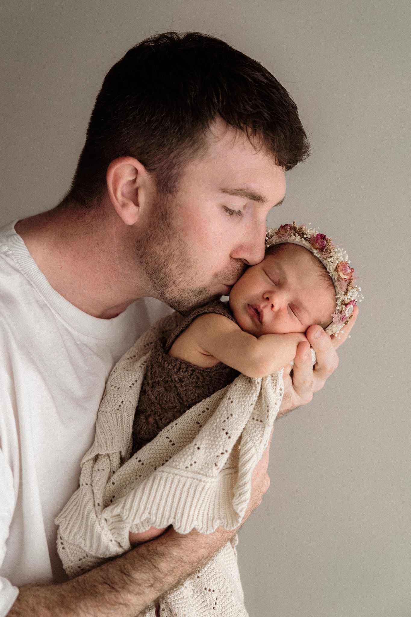 A tender moment shows a father gently kissing his sleeping newborn baby wrapped in a cream knit blanket.
