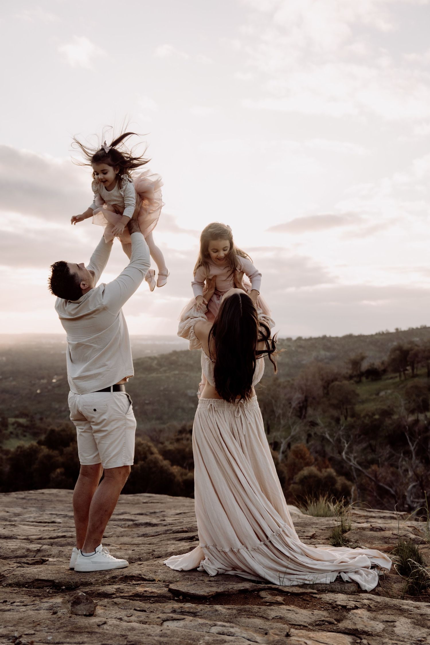 Family sharing a joyful moment together on a scenic cliff overlook at sunset, wearing flowing white outfits.