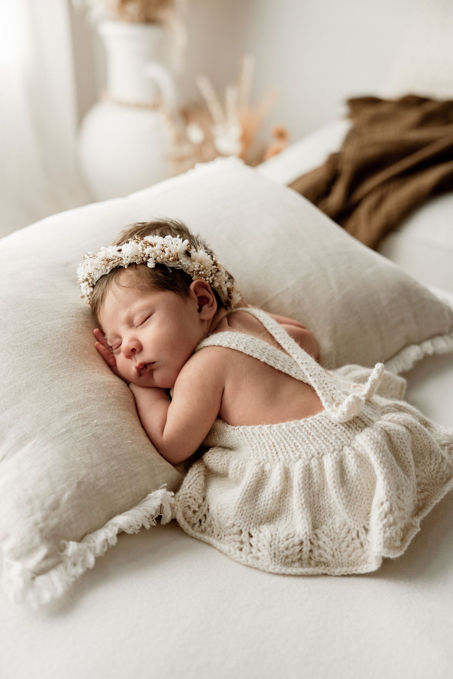 Sleeping newborn baby girl wearing a floral crown and cream knit romper resting on a white linen pillow.