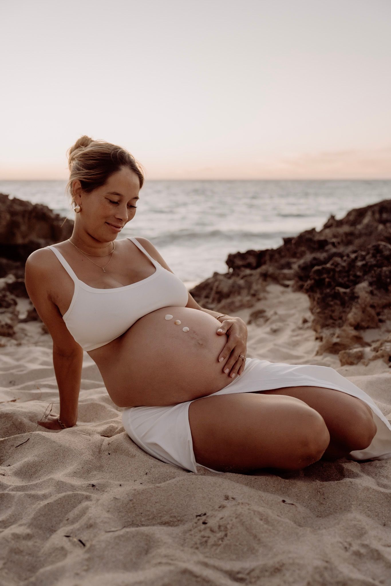 Pregnant woman in white swimwear rests on sandy beach at sunset with ocean and rocks in background.