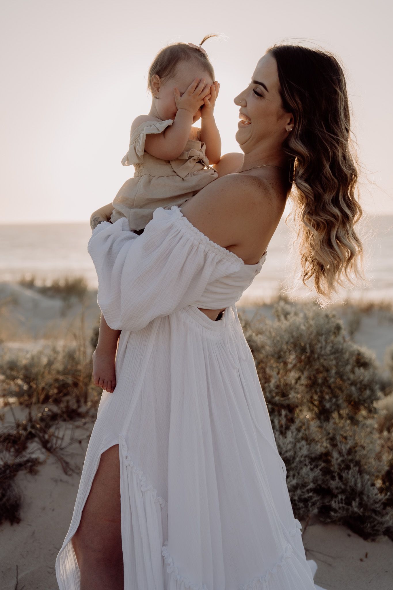 Woman in flowing white dress holds small child on sandy beach at sunset with dreamy backlit glow.