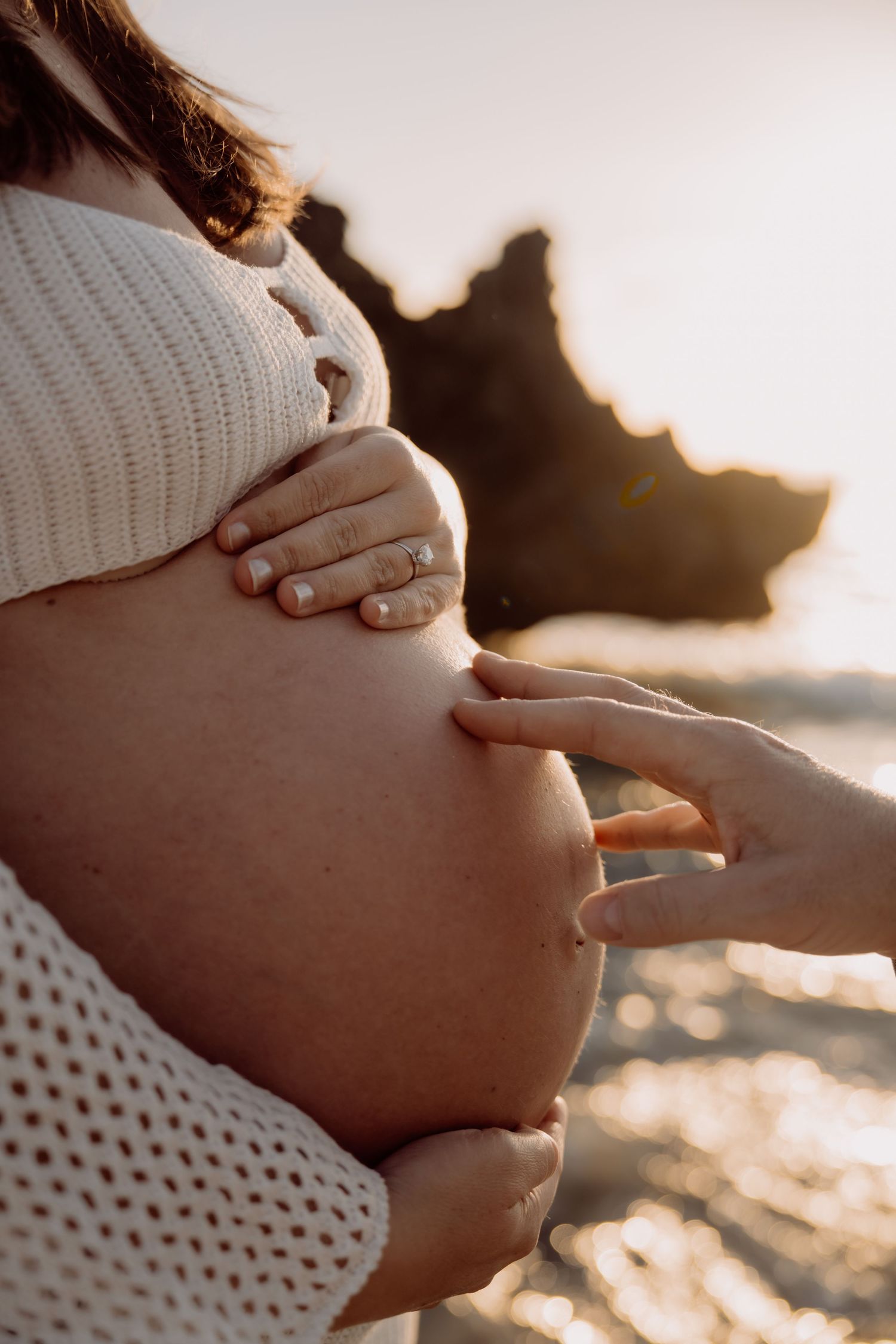 Pregnant woman in white knit top cradling her bare baby bump on a beach at golden sunset.