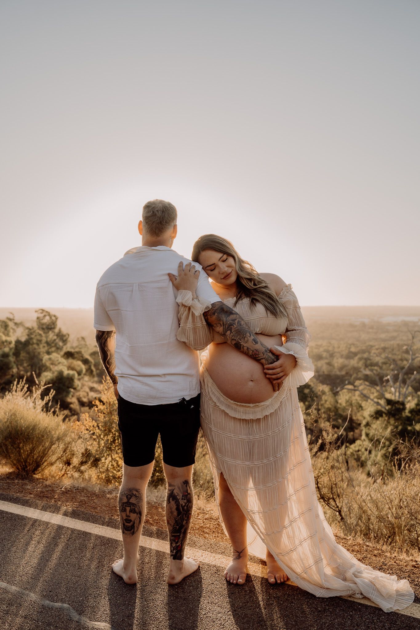 A couple stands together on a road at sunset, celebrating a maternity photoshoot with the countryside in the background.