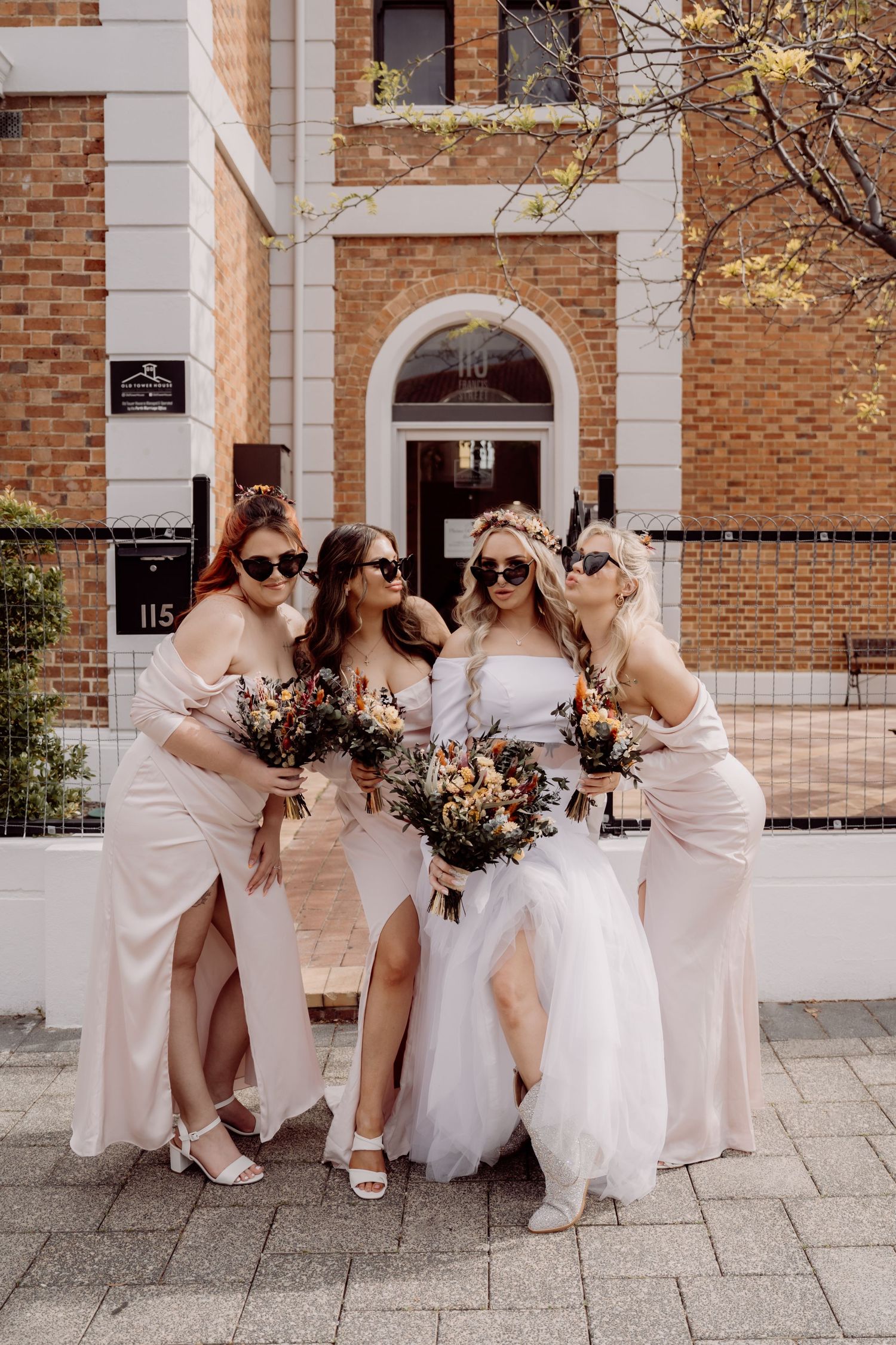 Bridesmaids in blush pink dresses and white gowns gather outside a brick building for candid wedding photos.