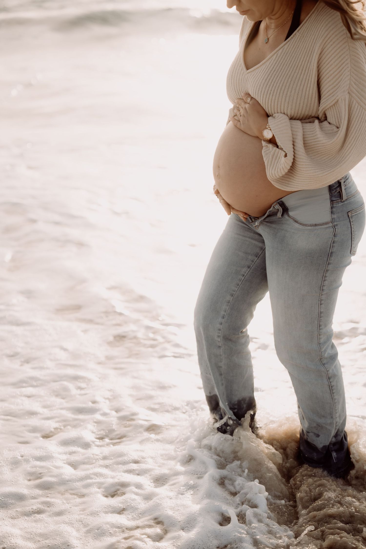 Pregnant woman in beige sweater and jeans cradling baby bump while standing in ocean waves on a beach.