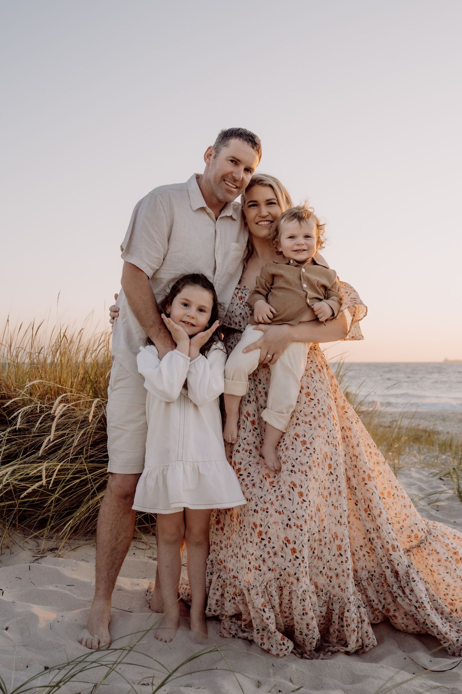 A family sits together on a sandy beach at sunset wearing coordinated white and floral outfits.