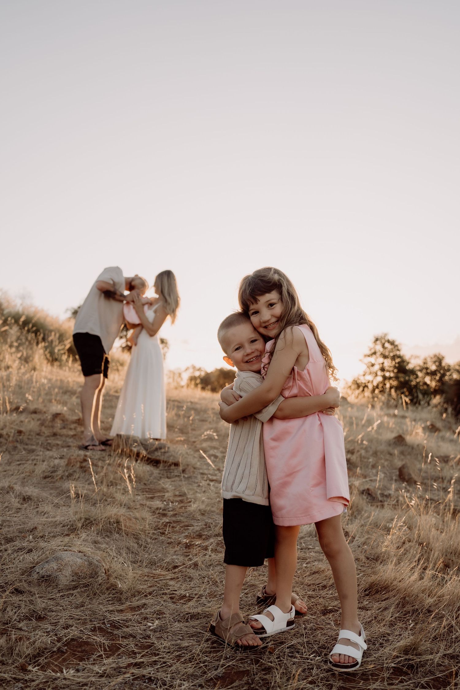 A loving family sharing embraces together on a hillside at sunset in dry grass against warm golden light.