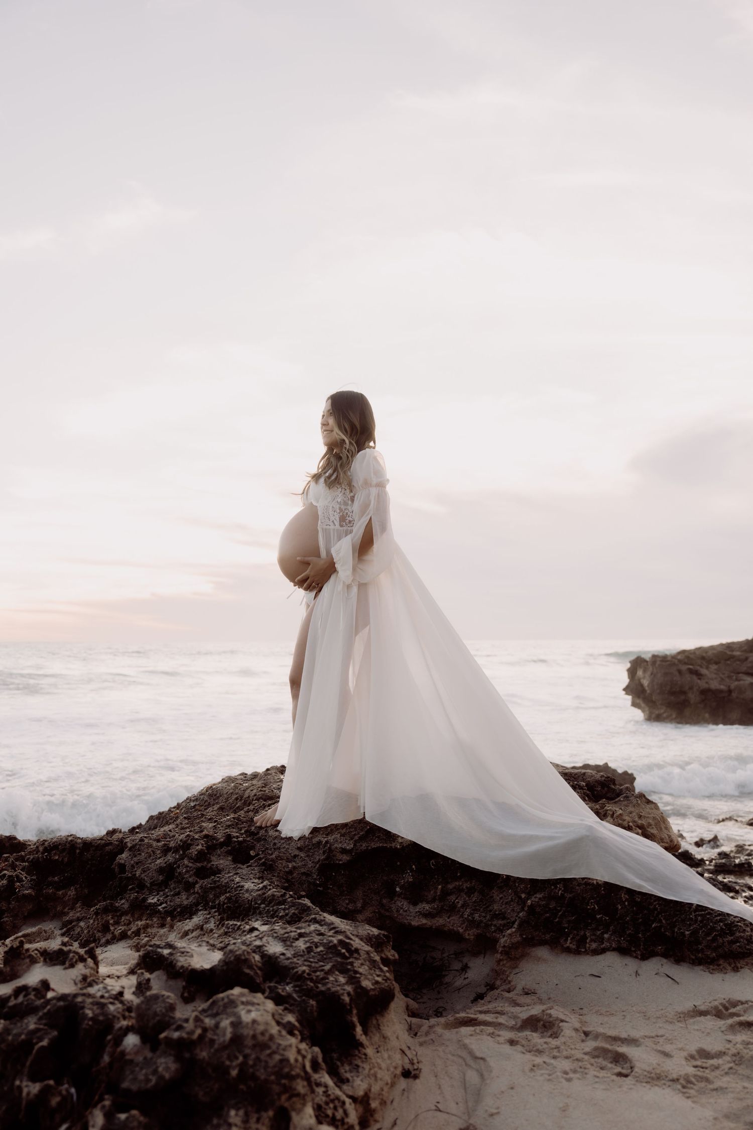 Pregnant woman in flowing white gown standing on rocky beach at sunset, cradling baby bump while ocean waves crash behind her.